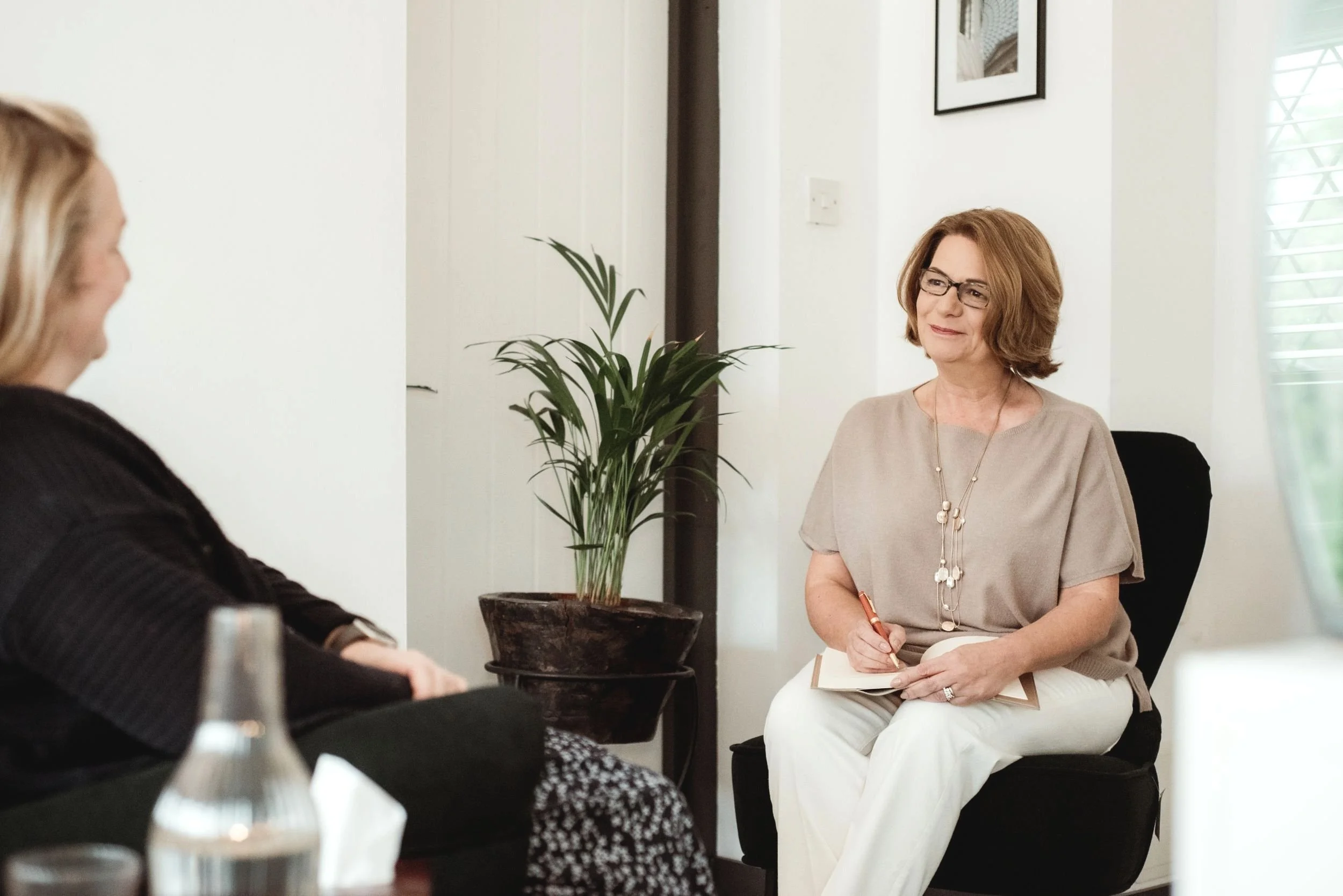 Client in a black top speaking on the left, while therapist Lois on the right listens and holds a pen poised over a notebook, both seated