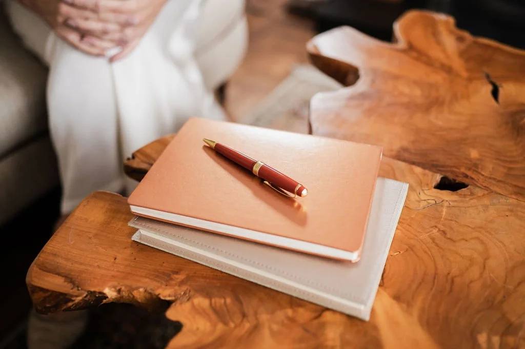 Pen resting on two stacked notebooks on a wooden coffee table, with legs of a seated person visible in the background