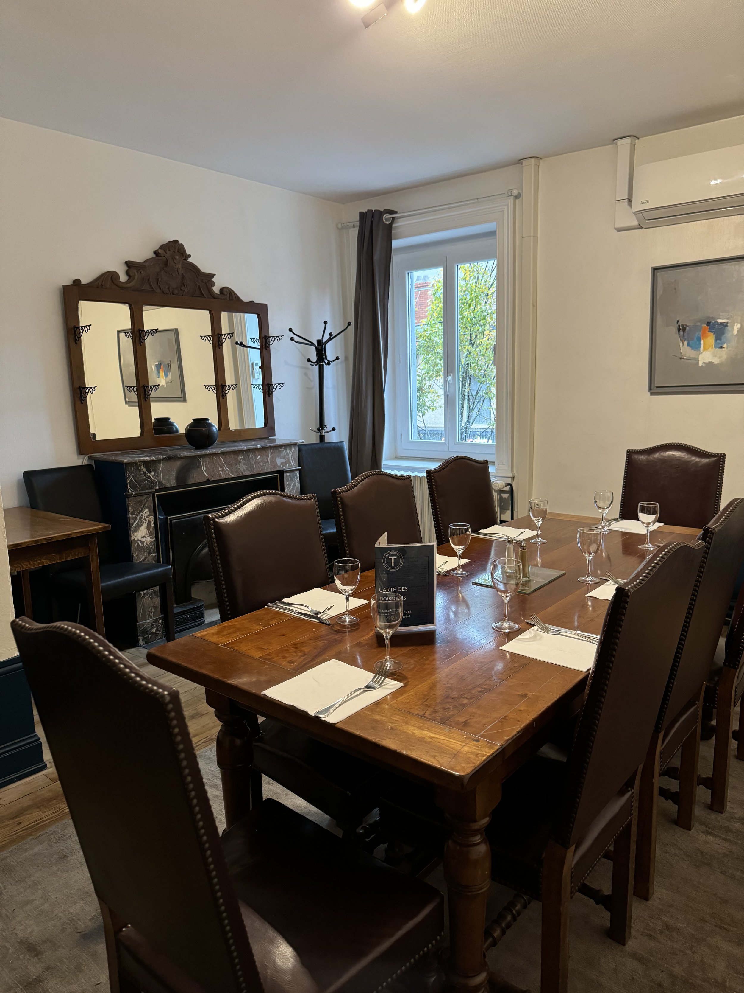 Une salle à manger ou de réunion avec une grande table en bois entourée de huit chaises en cuir, un miroir ancien sur cheminée en marbre, une fenêtre avec rideaux, une œuvre d'art abstrait sur le mur, et une lampe au plafond, comme à la maison.