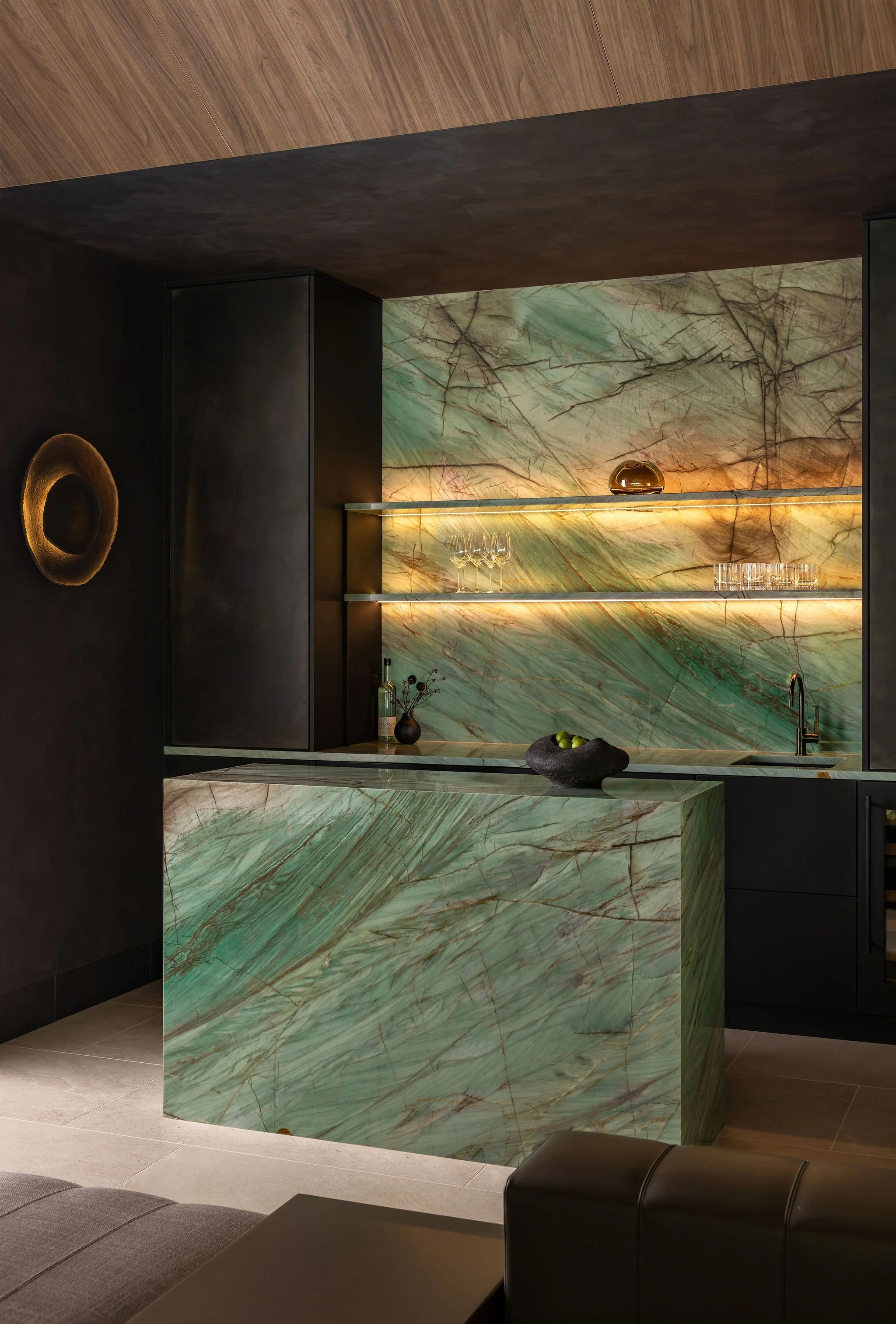 Modern, moody dark bar with lit stone shelves and dark cabinetry and full stone island