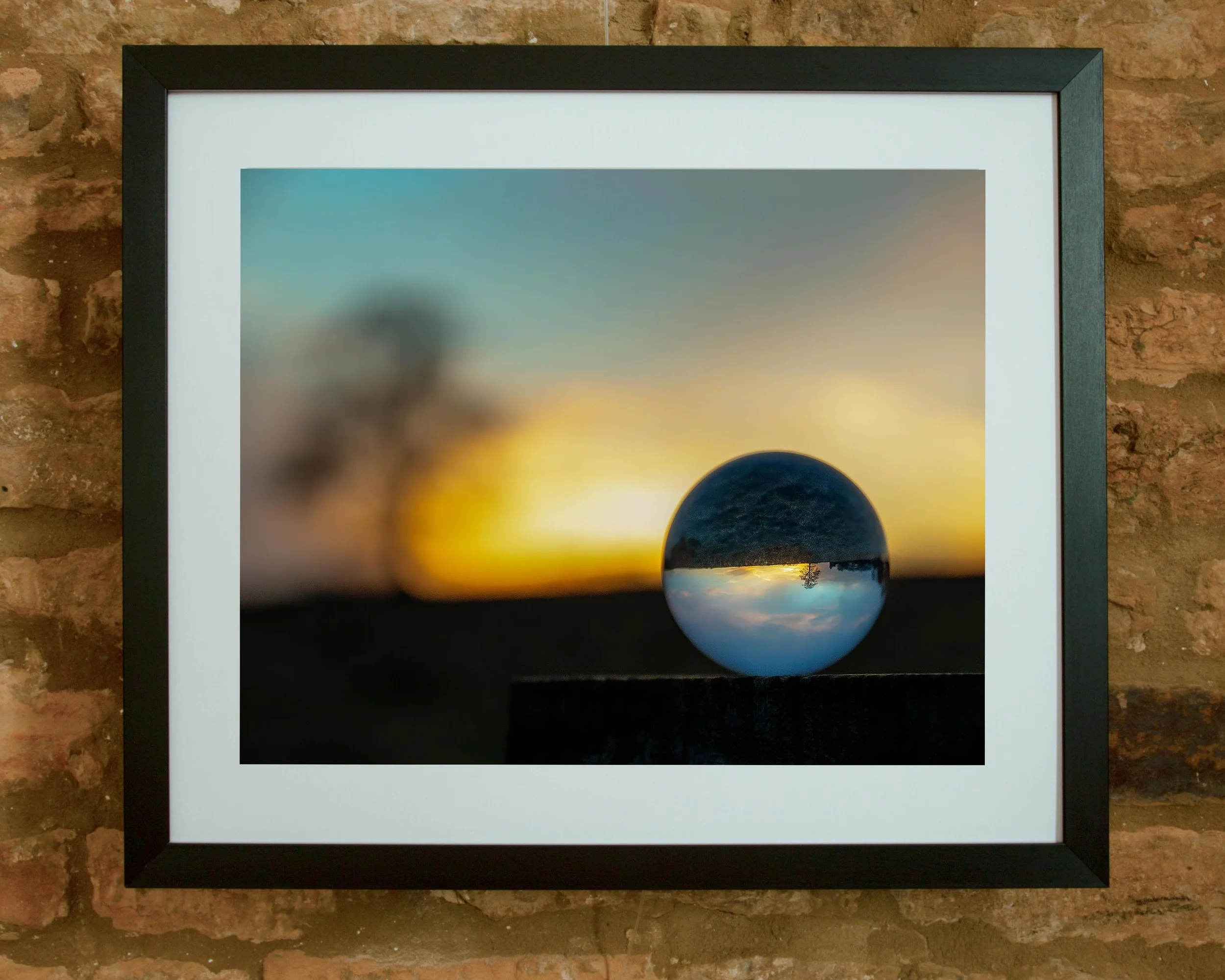A framed photograph of a sunset with a blue and white marble ball in front of it, placed on a brick surface.