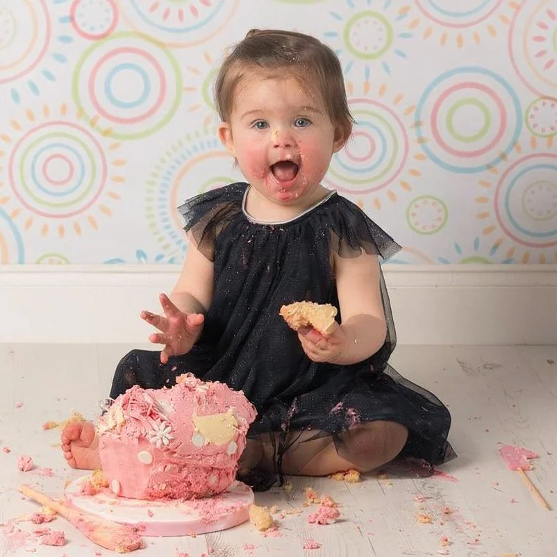 Child sitting in blue dress against patterned backdrop, cake smash photography