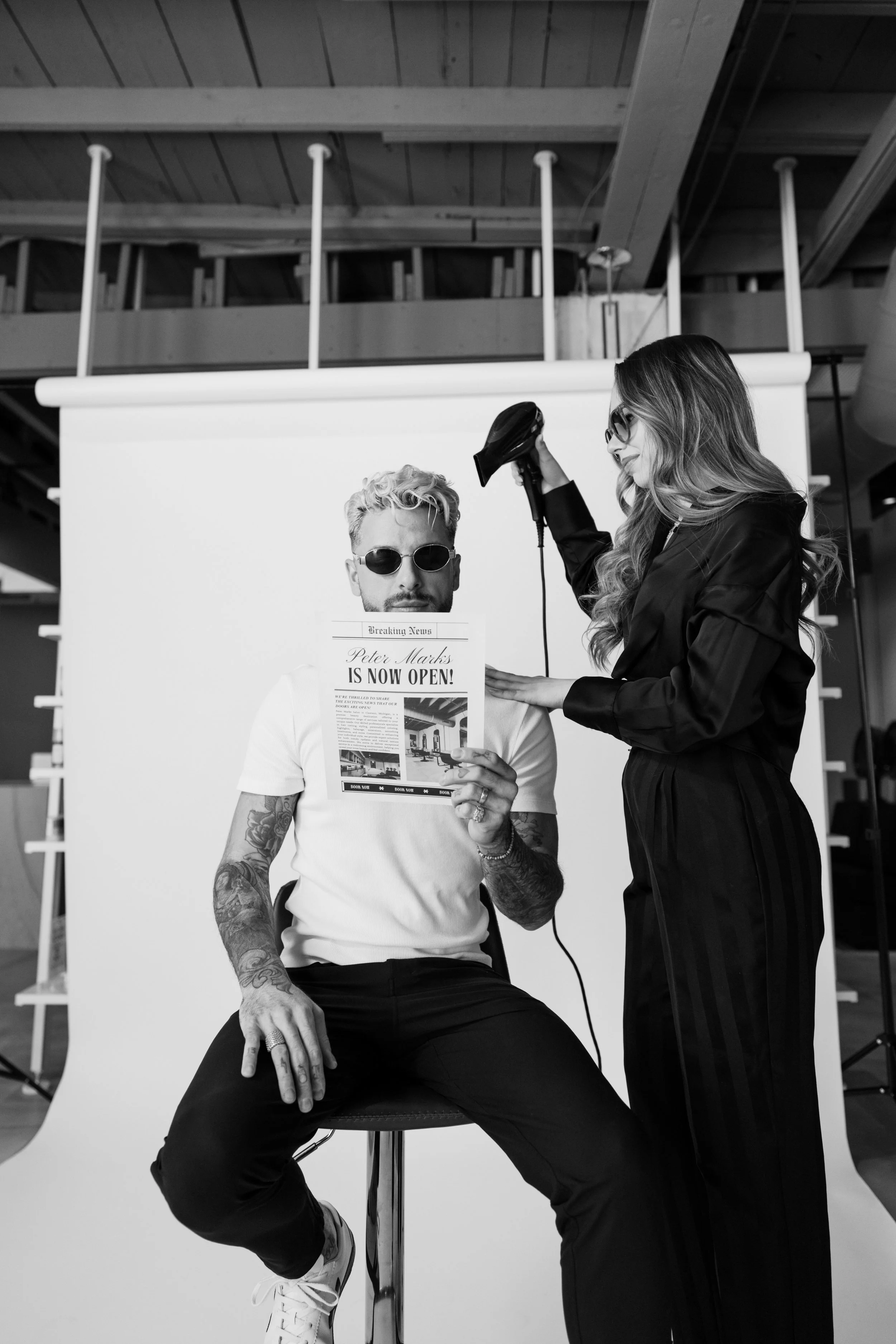 A person in sunglasses sitting on a stool holding a flyer or newspaper headline that reads "Peter Marks Is Now Open!" A hair stylist stands next to them using a hairdryer. The setting is in Peter Marks Salon.