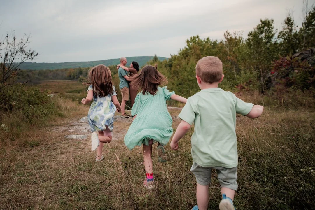 Four children running down a trail in a natural, grassy outdoor area with trees and hills in the background.