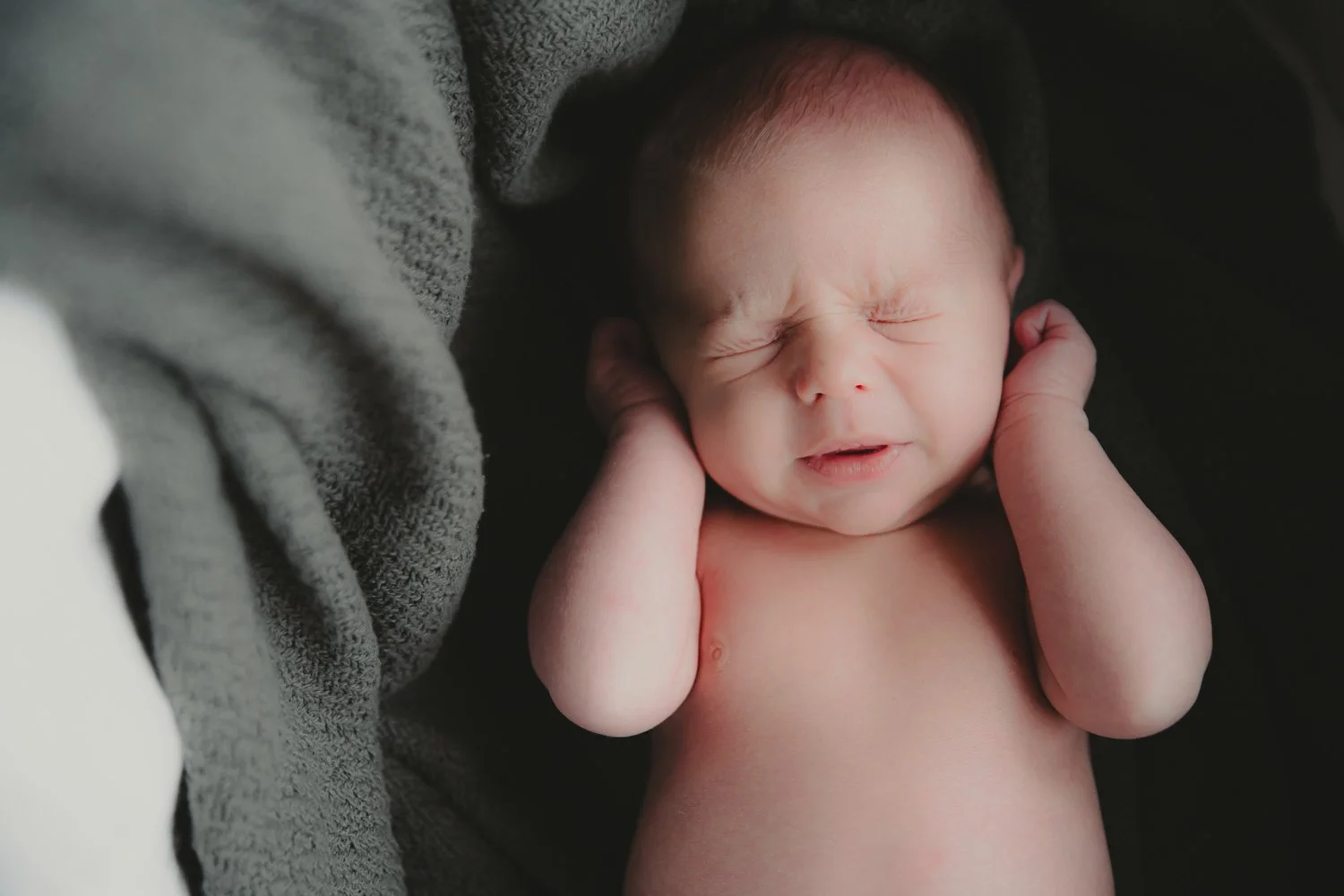 A newborn baby is holding its ears with both hands, with a scrunched face and closed eyes, lying on a dark surface.