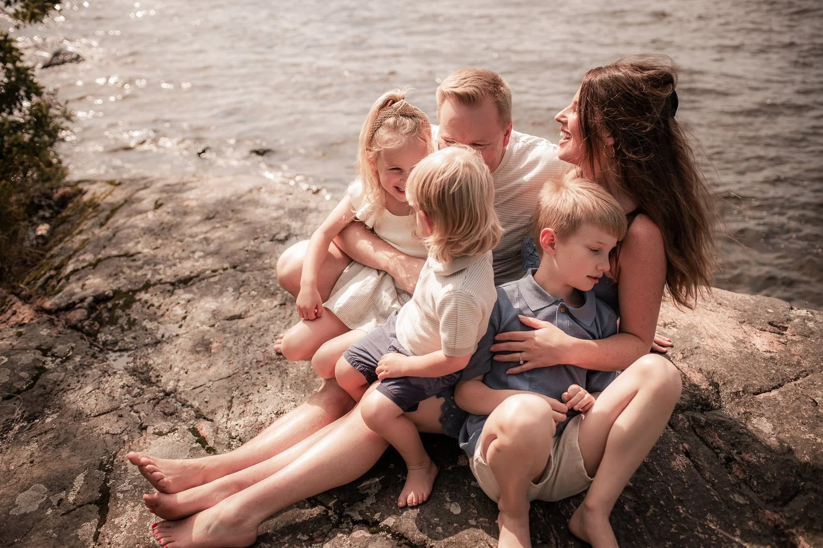 A family of five sitting on rocks near a body of water, enjoying a sunny day outdoors.