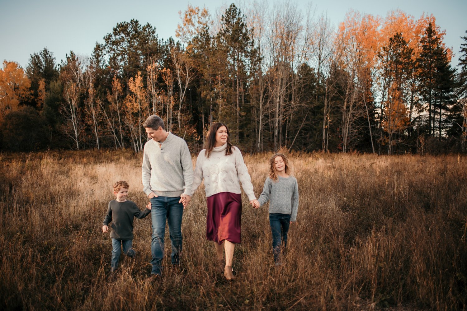 A family of four walking through a grassy field during fall, holding hands, with trees with autumn foliage in the background.
