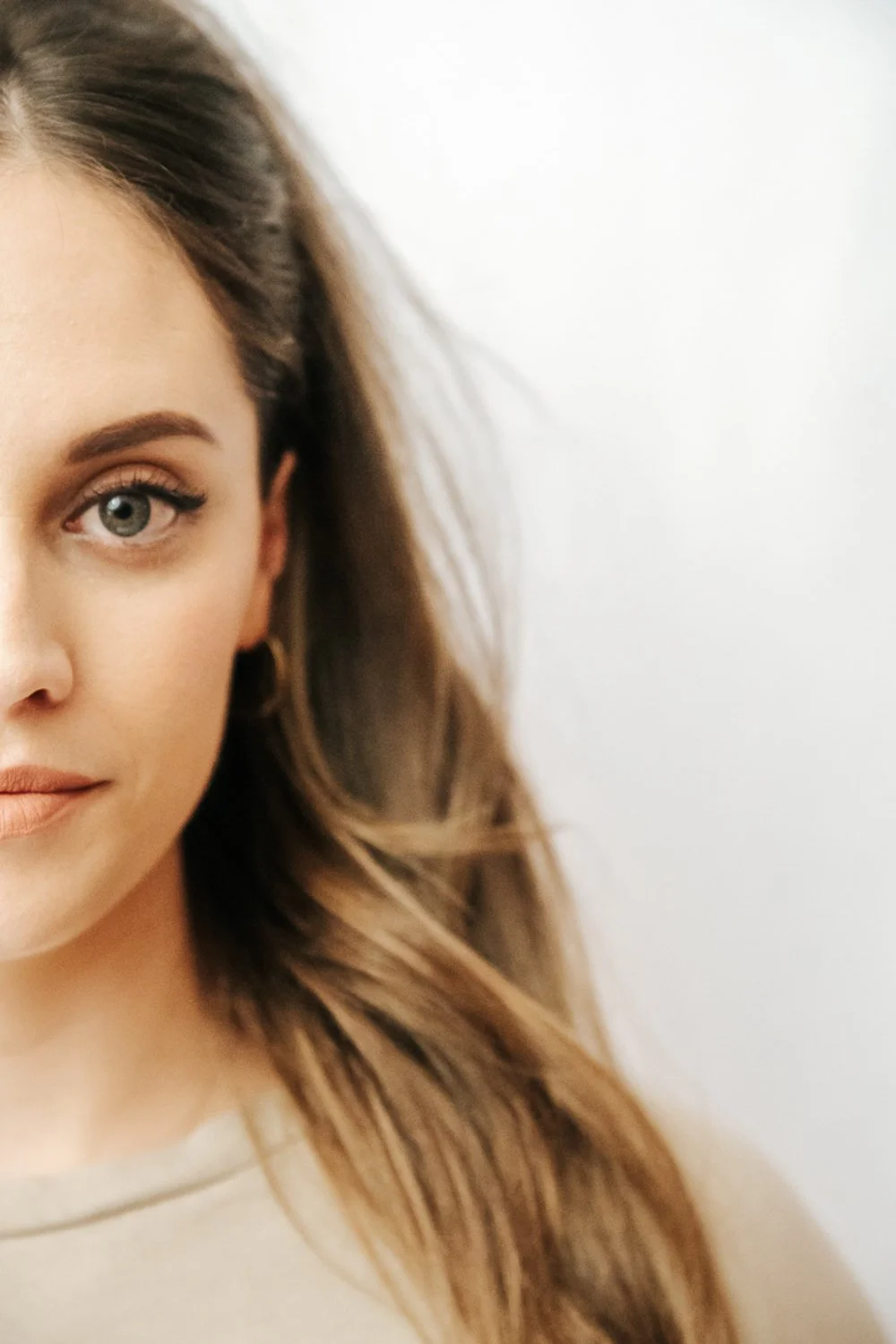 Close-up of a woman's face showing her left eye, part of her nose, lips, and flowing brown hair against a plain white background. Eye Lash Technician. 