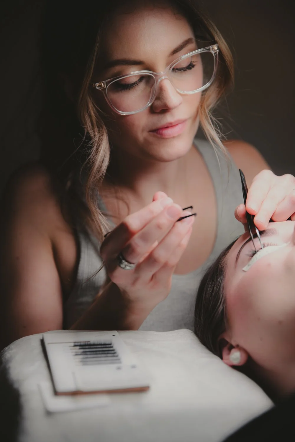 A female cosmetologist performs eyelash extensions on a client lying down, wearing protective glasses, with equipment on a table in front.