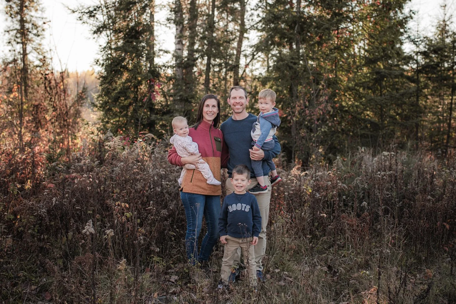 A family of five stood outdoors in a wooded area during autumn. The parents, a man and a woman, each hold a child, and there is a young boy standing in front. The family is smiling and dressed in casual clothing suitable for cool weather.