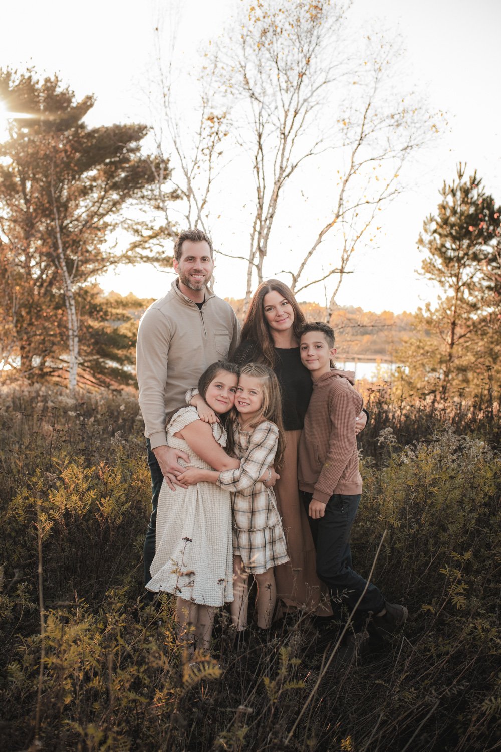 Family of five standing outdoors in autumn, smiling, with trees and a lake in the background.