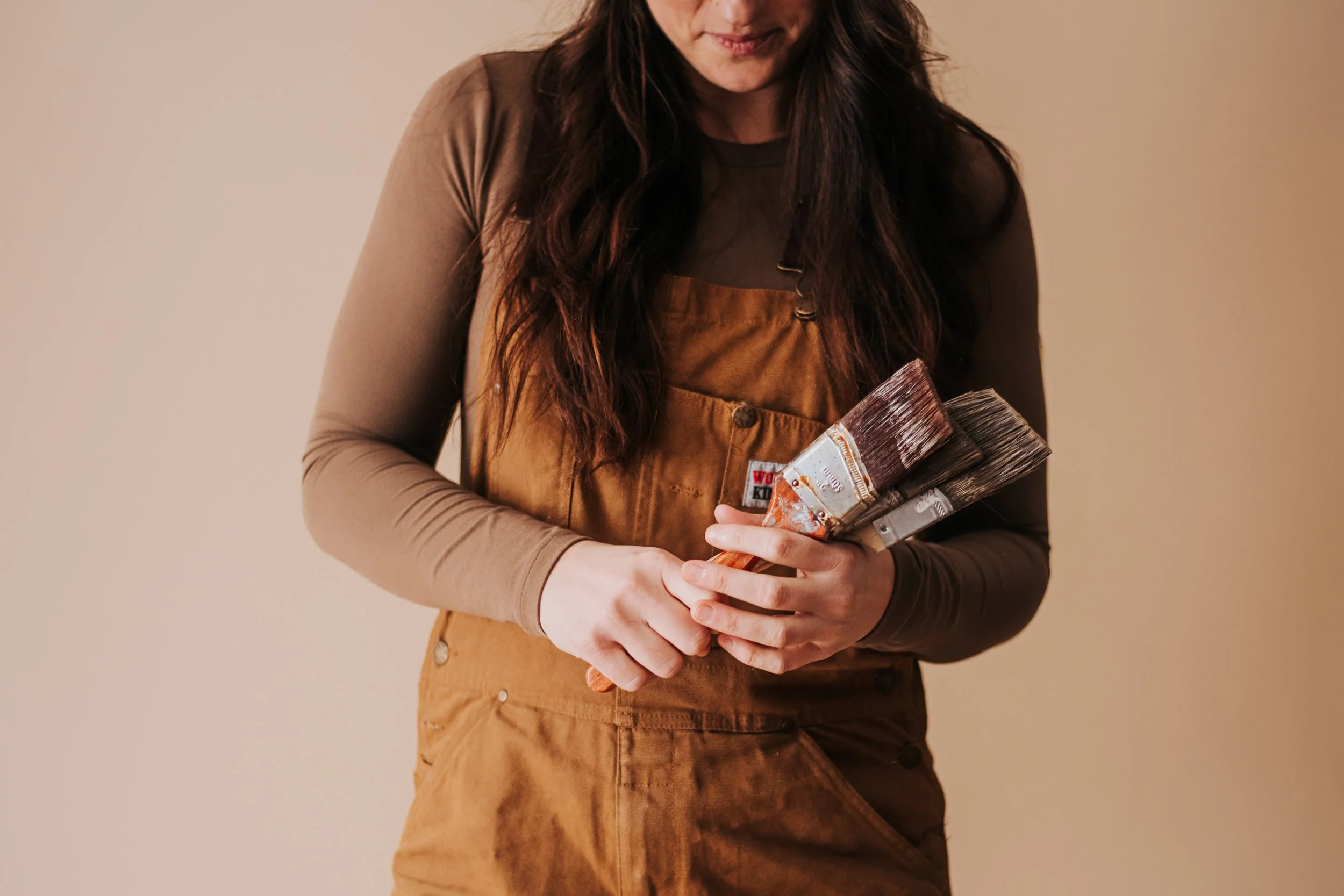 Person holding several paintbrushes, wearing a brown apron and a long-sleeve shirt, against a plain background.