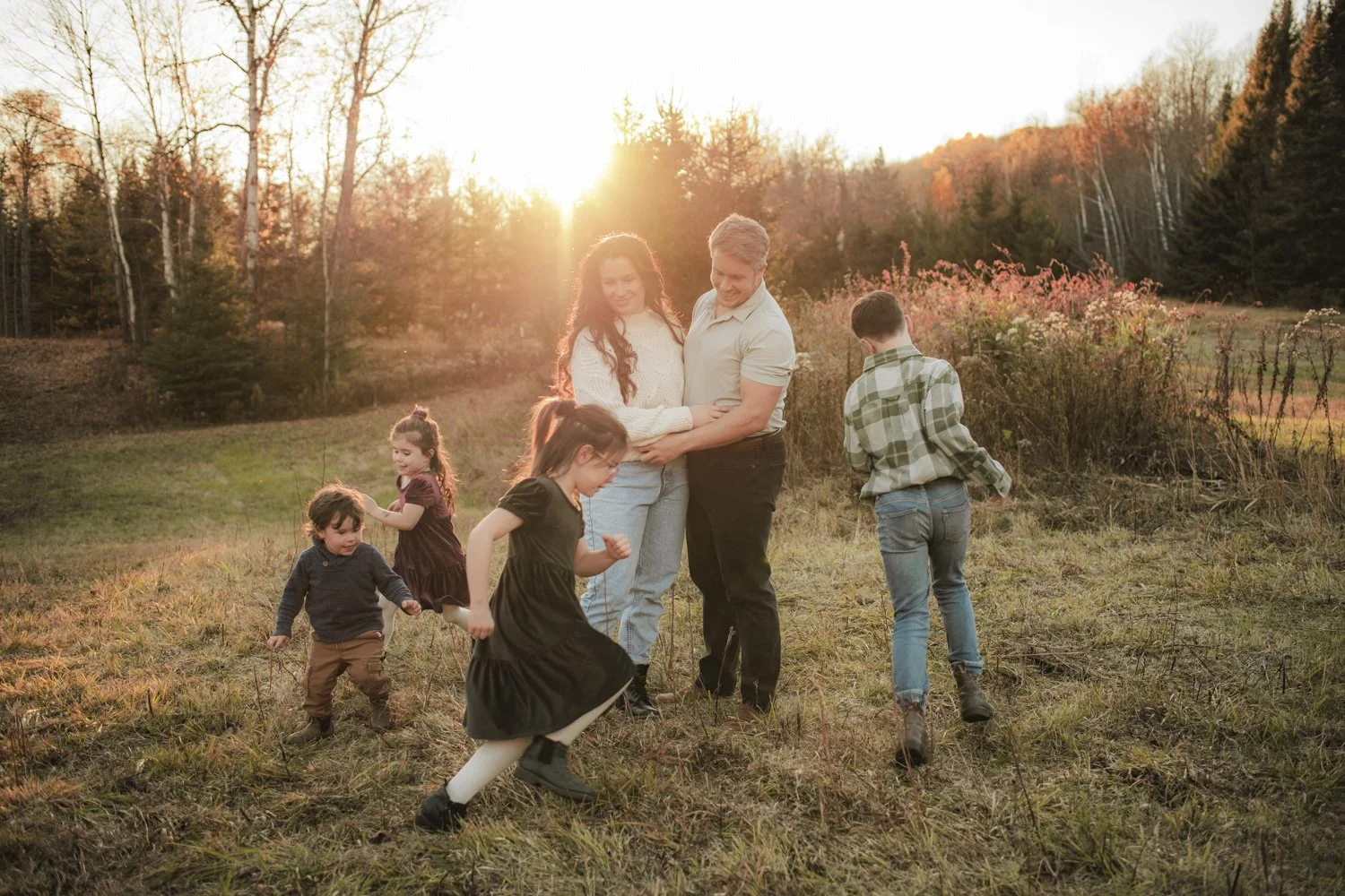 Family of six children and two adults, including two girls, one boy, and two younger children, standing and playing outdoors in a grassy field during sunset, with trees and hills in the background.