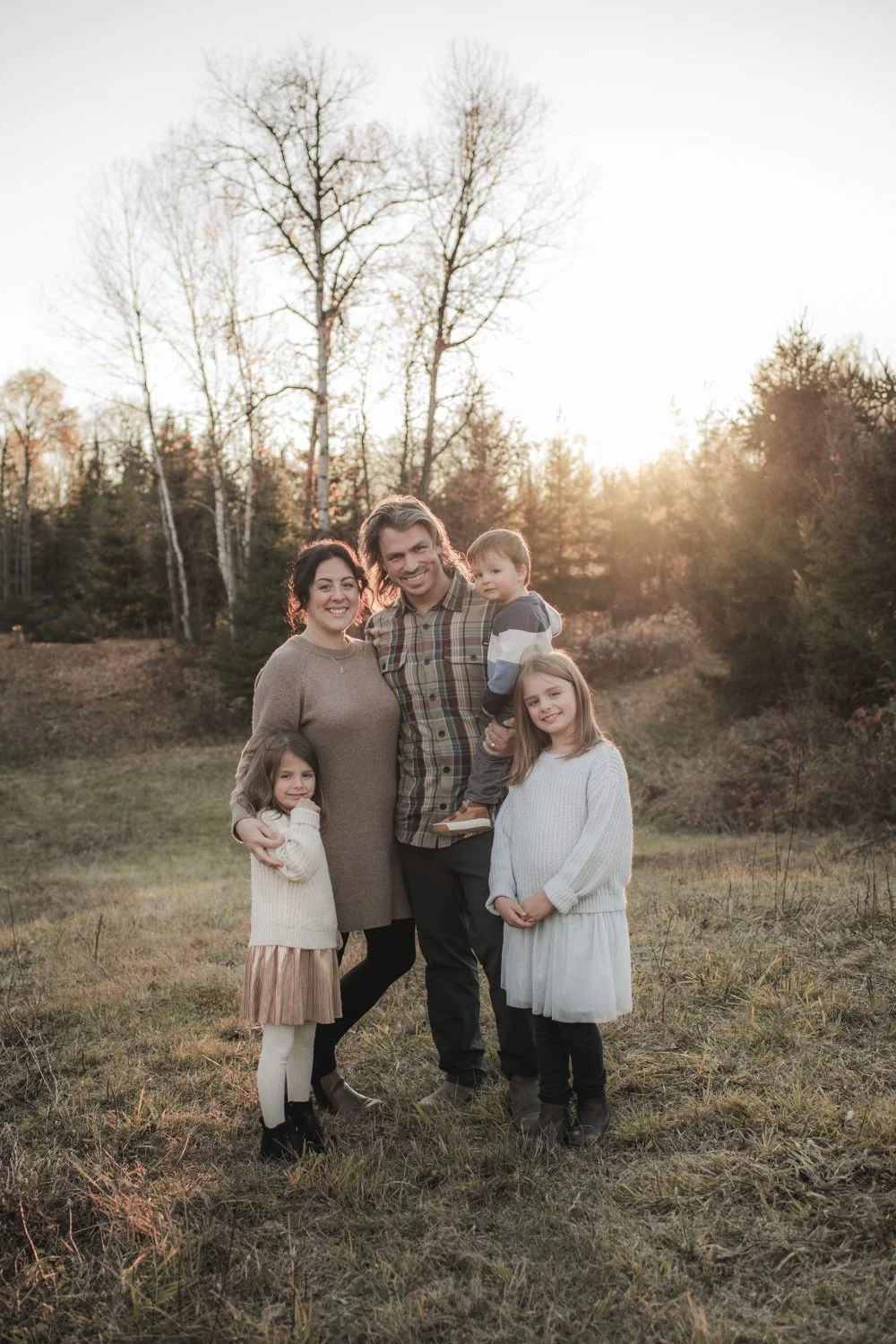 A family of five standing outdoors in a field at sunset, smiling for a photo. The family includes a man, a woman, and three children. The background has trees and a bright sky.