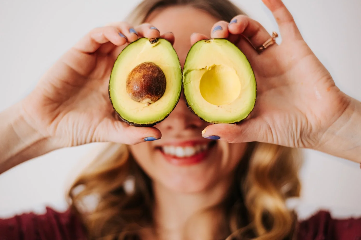 A woman holding a halved avocado in front of her face, with one half showing the seed and the other half empty, smiling and blurred in the background.