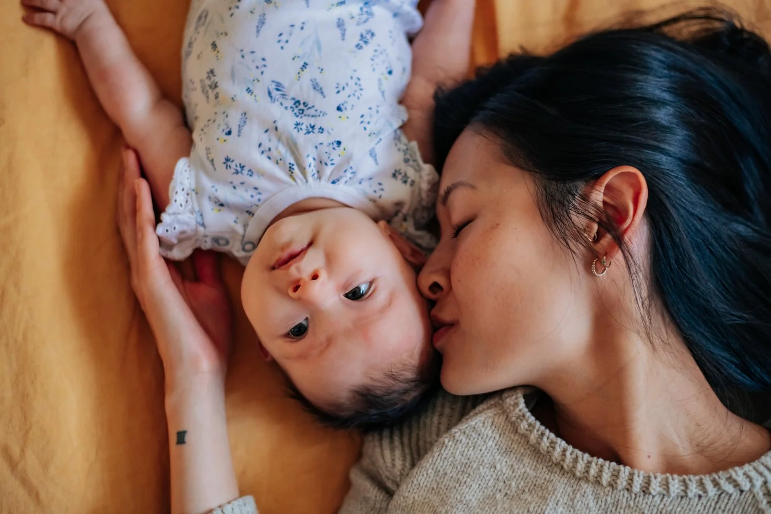 A woman with dark hair and earrings is lying down on a wooden surface, gently kissing a baby with light skin, blue eyes, and short dark hair who is lying on her side, wearing a white onesie with purple and yellow floral patterns.