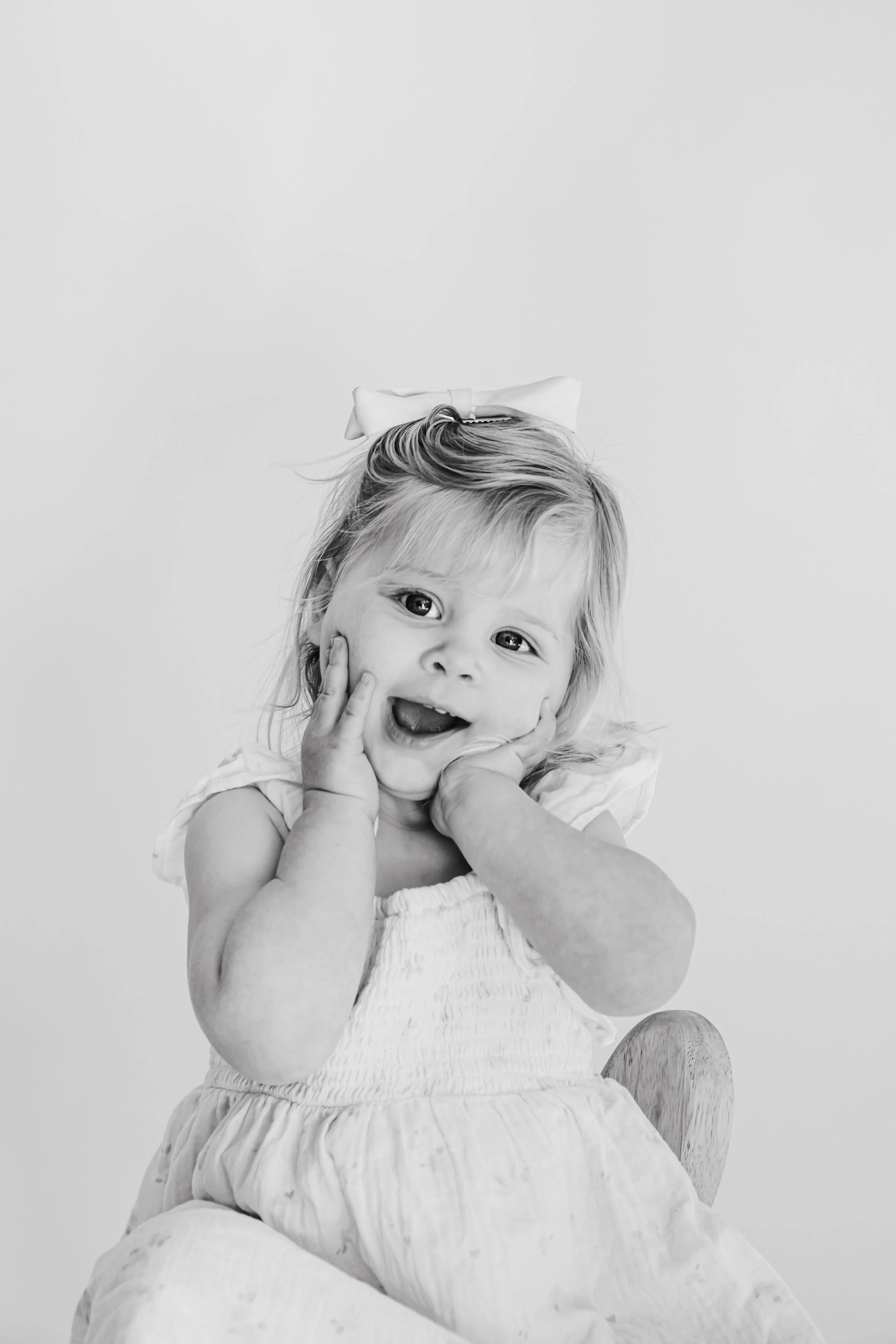 Black and white photo of a young girl with a big white bow in her hair, smiling and holding her face with both hands, sitting on a wooden chair.