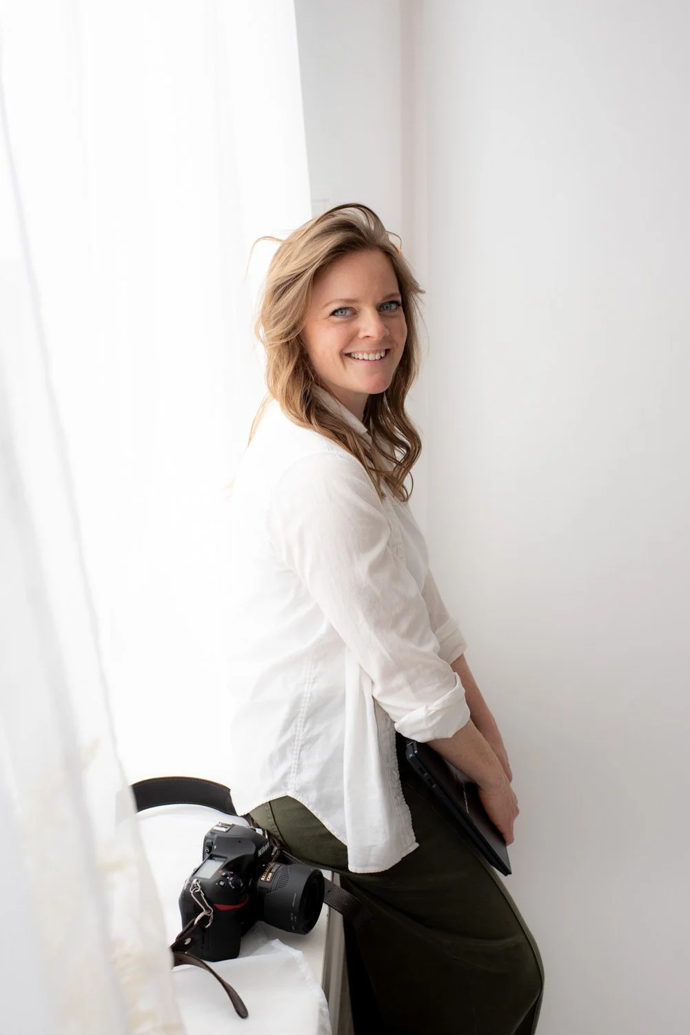 Smiling woman sitting on a chair near a window with sheer white curtains, holding a black laptop, with a camera placed on a white surface nearby.