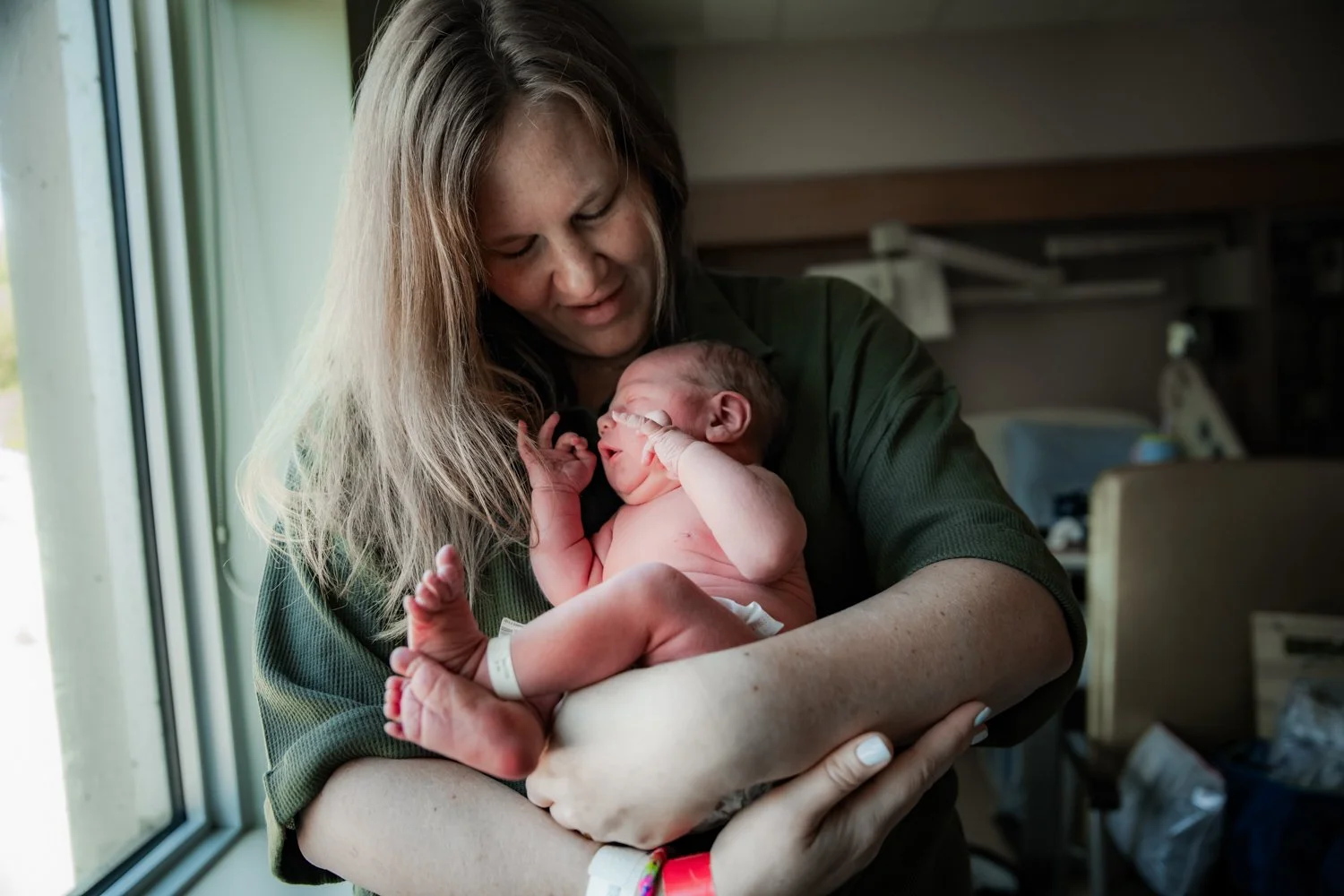 A woman holding a newborn baby in a hospital room, smiling and looking down at the baby, who is lying on her arms.