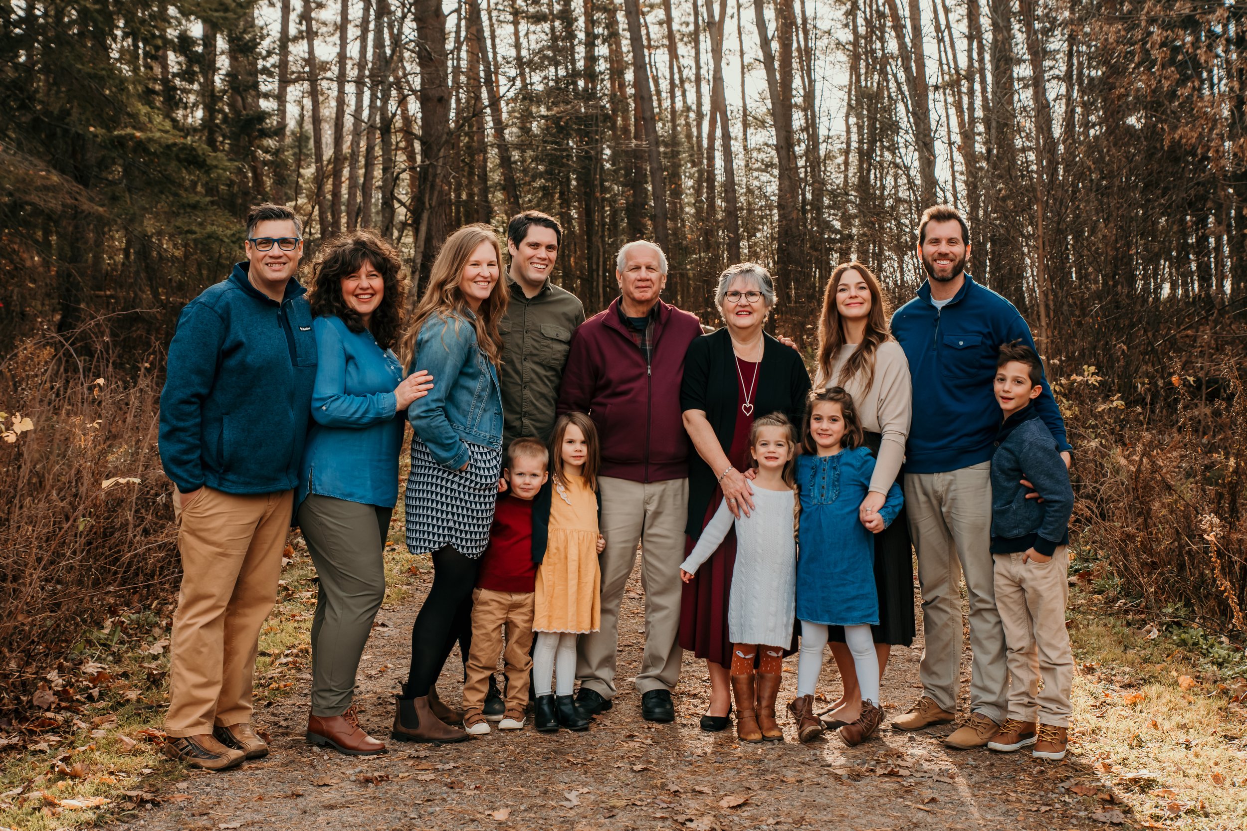 A family group of ten people, including children, standing on a wooded trail during autumn, smiling at the camera.