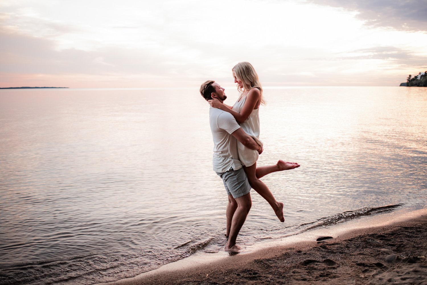 A couple is standing on the beach, with one person lifting the other off the ground, in front of a calm body of water during sunset.