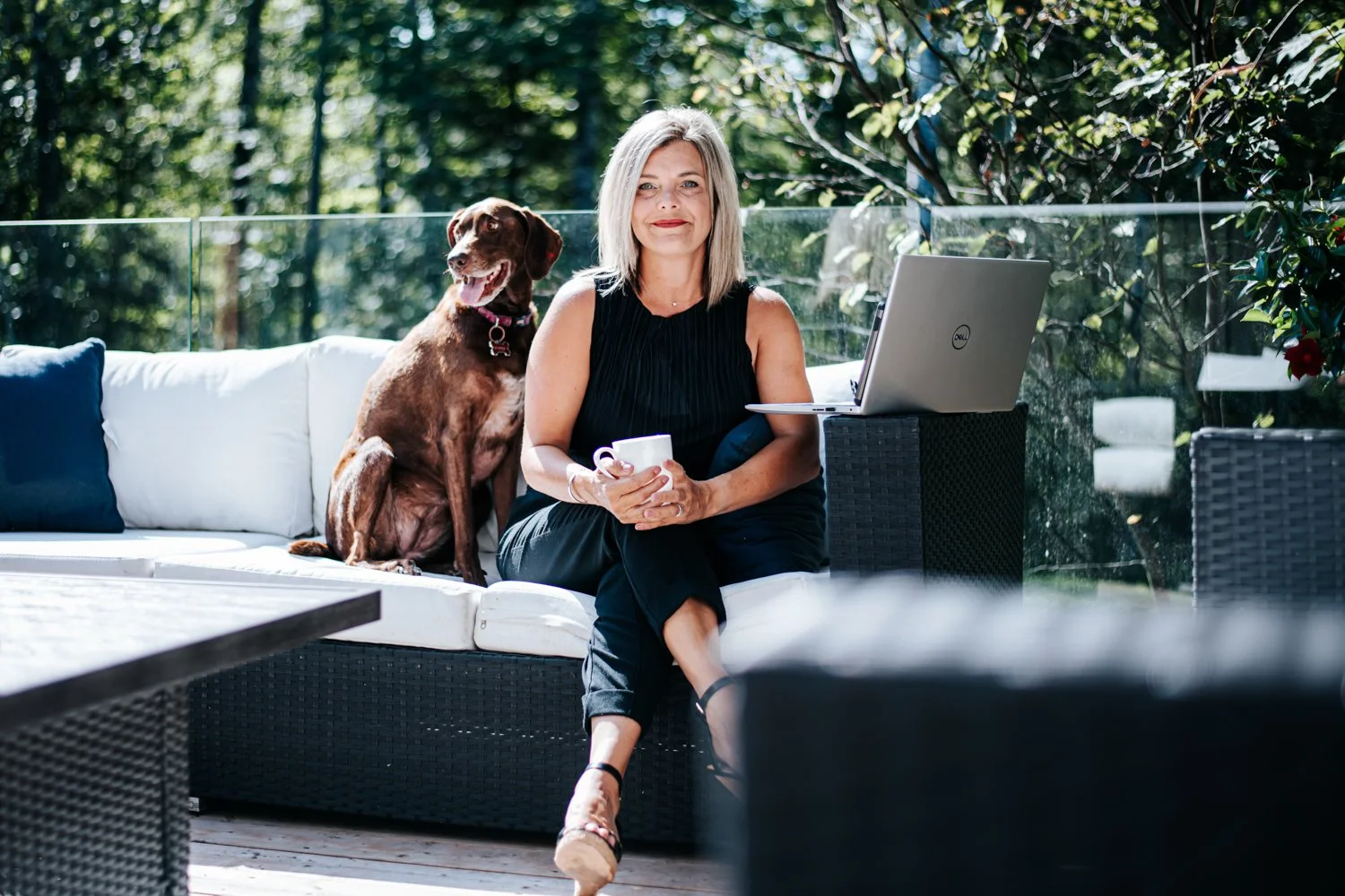 A woman sitting outdoors on a white couch with a brown dog beside her, holding a mug, with a laptop on a black stand in front of her.