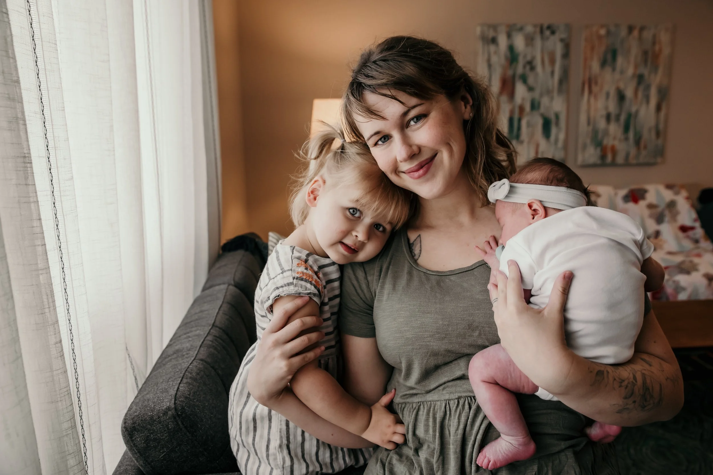 A woman holding a newborn baby with a headband, and a young girl leaning on her shoulder inside a living room.