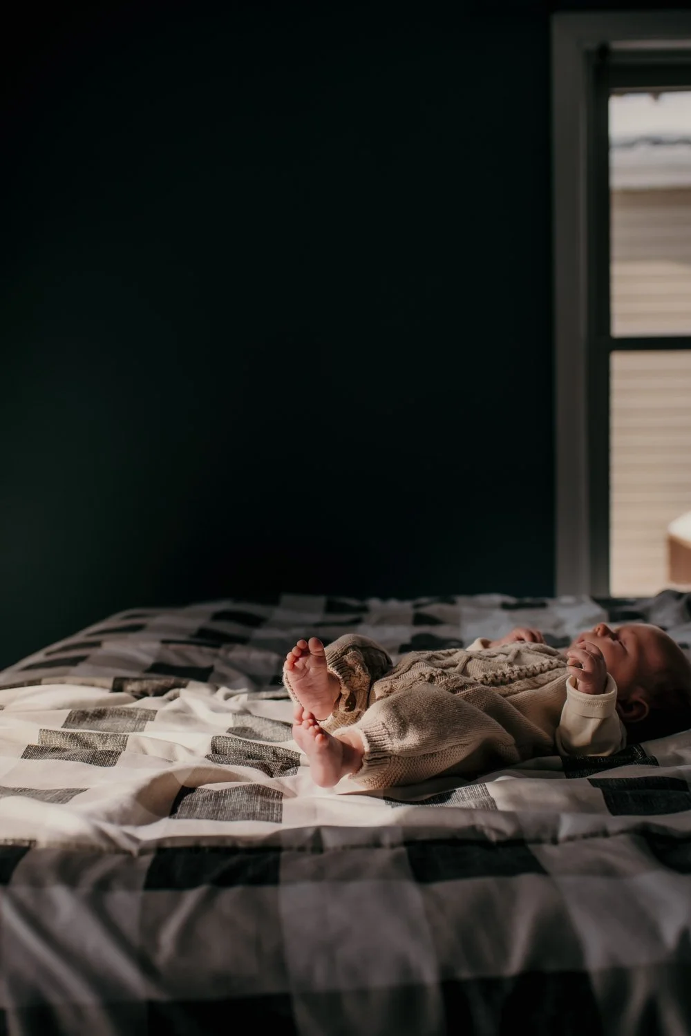 A baby lying on a bed with sunlight streaming through a window, wearing a knitted sweater and pants, with a happy expression.
