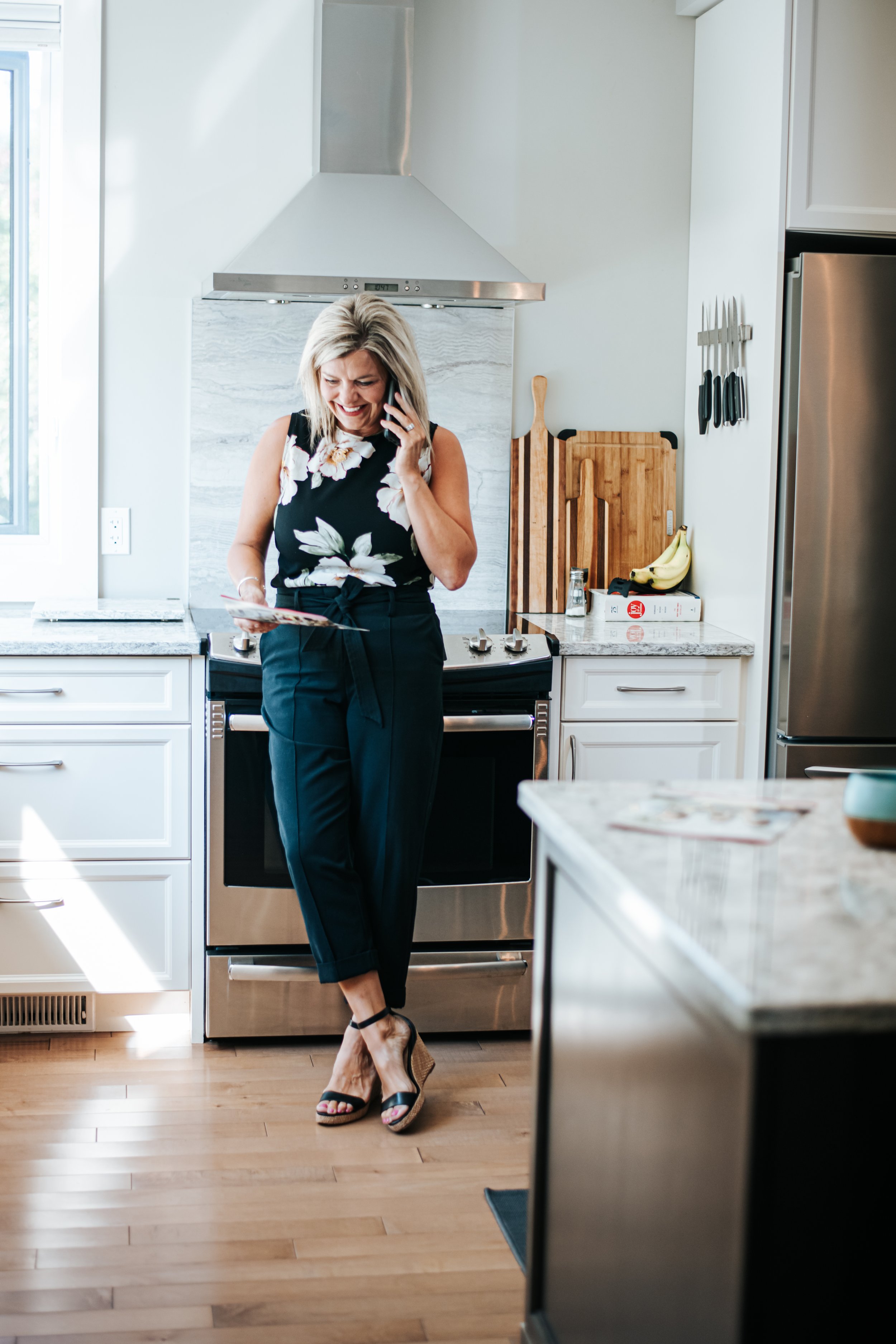 A woman in black pants and a floral top standing in a modern kitchen, talking on her phone and holding a magazine. Real estate Agent
