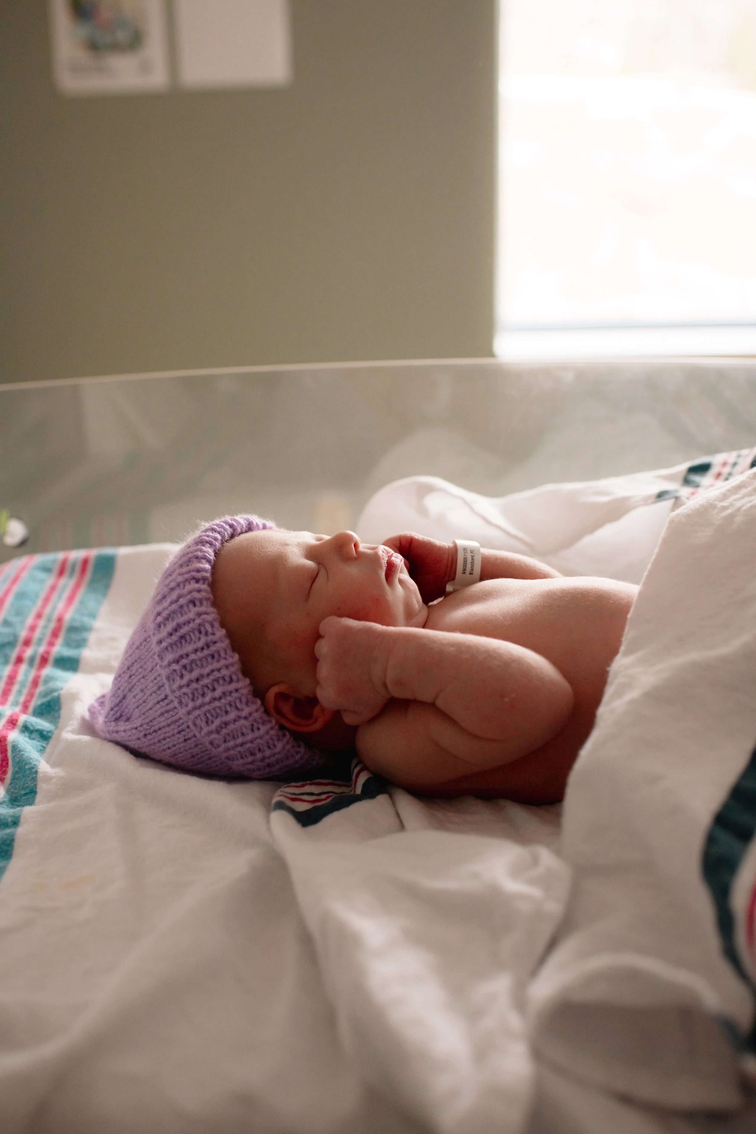 A newborn baby lying on a hospital bed, wearing a purple knitted hat, with arms crossed near face, and a hospital wristband.