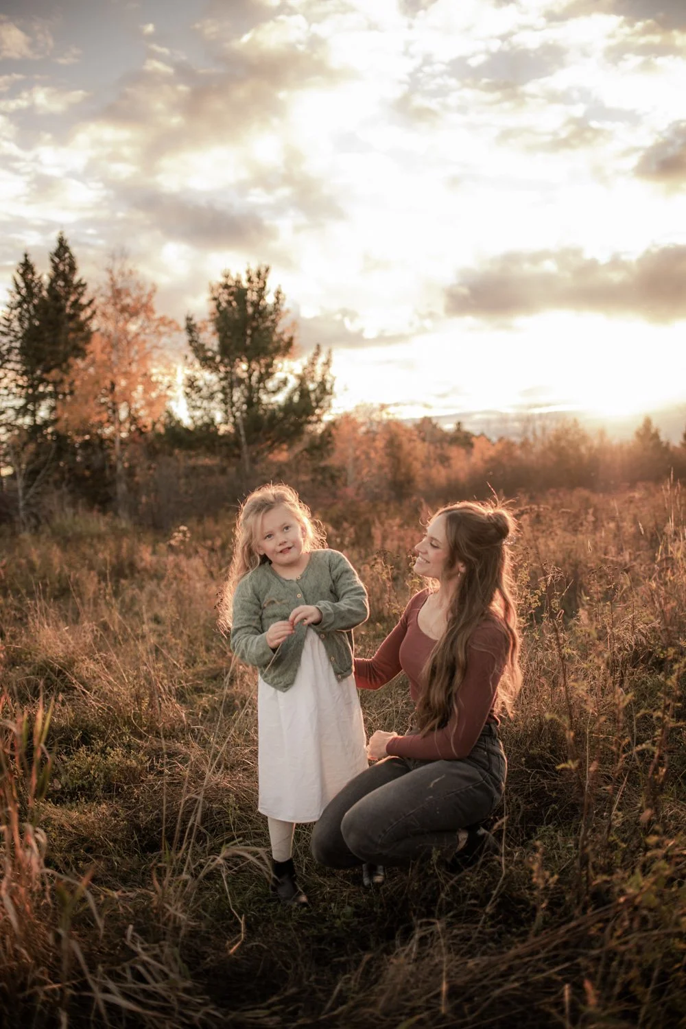 A woman and a young girl in a field during sunset, with the woman kneeling and smiling at the girl, who is standing and looking at the camera.