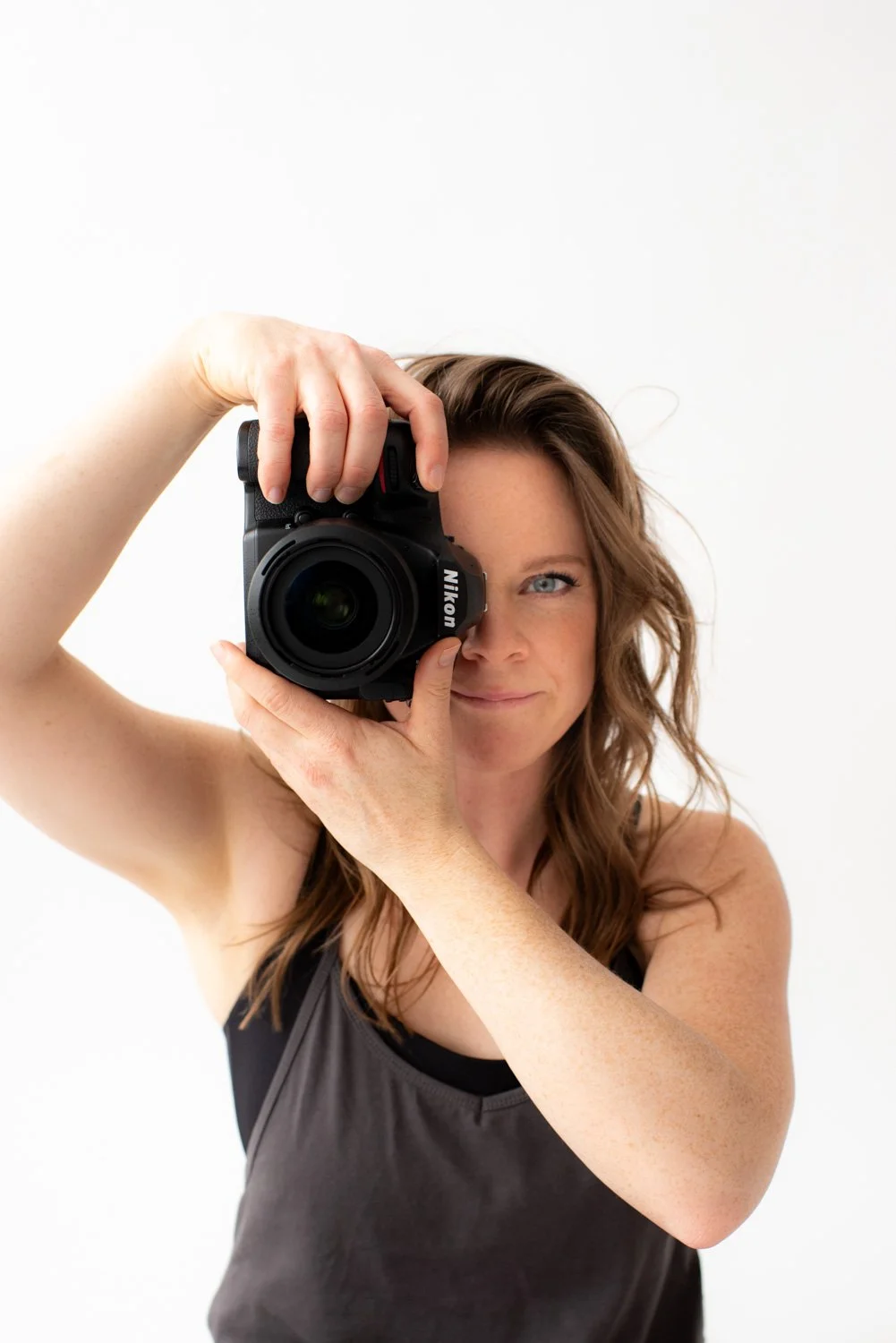 Woman with wavy brown hair taking a photo with a Nikon camera, smiling slightly, against a white background.