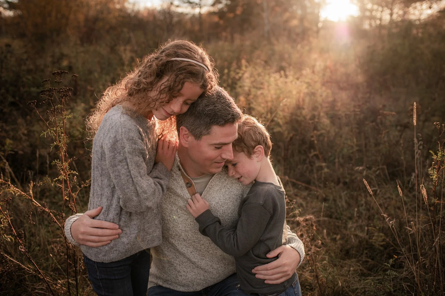 A family of three, a mother, father, and son, sharing a tender moment outdoors during sunset in a natural setting with tall grass and trees.