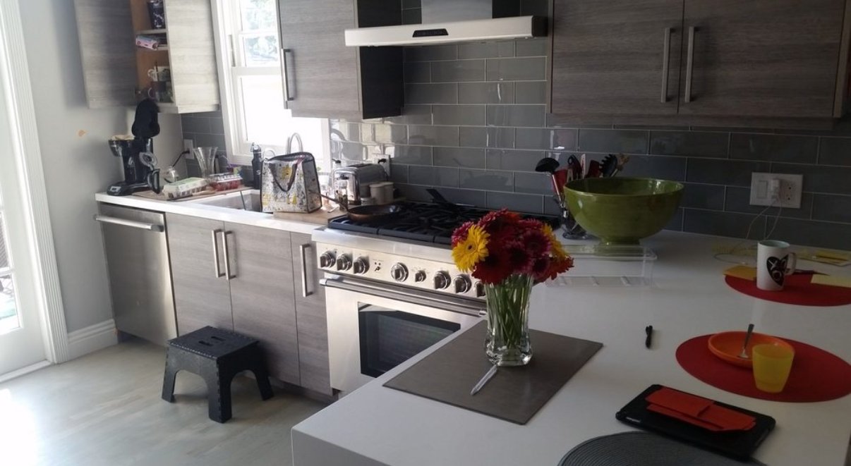 Modern kitchen with gray cabinets, a white countertop, a stove with a flower vase in the foreground, and various kitchen tools and appliances on the counter.