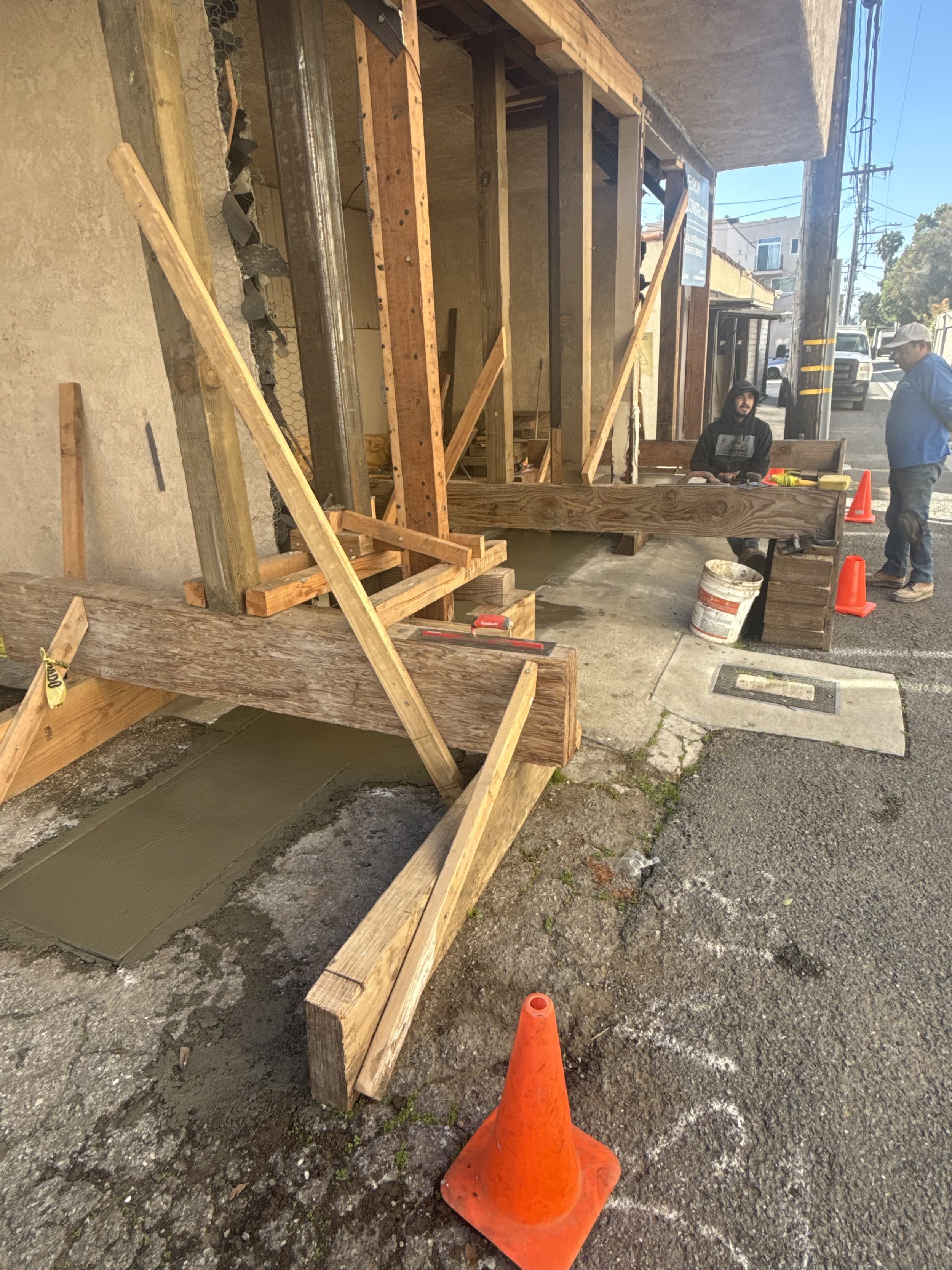 Construction site with wooden framing, workers, and orange traffic cone on the sidewalk.