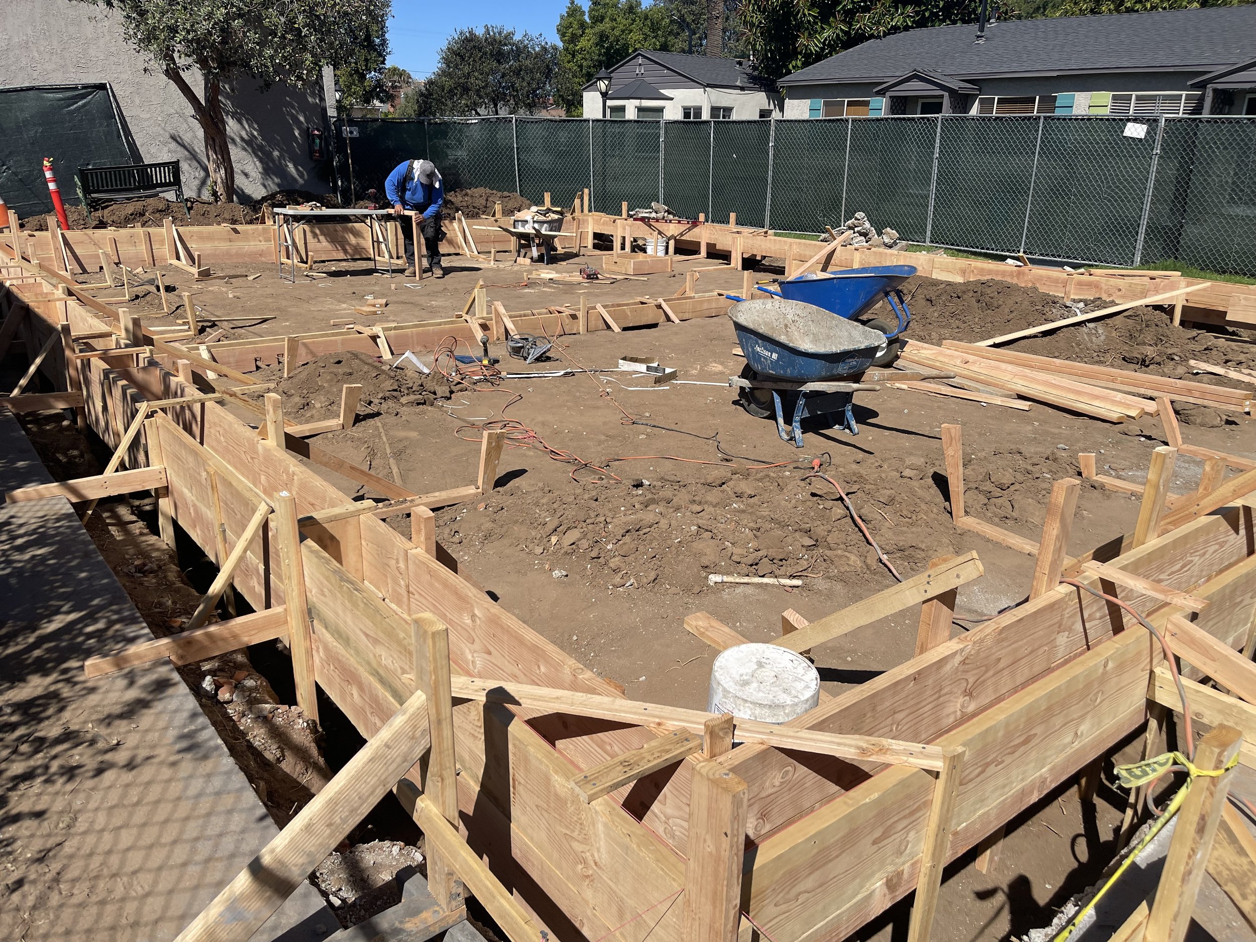 Construction site with wooden forms for a building foundation, two wheelbarrows, and workers in the background preparing the area.