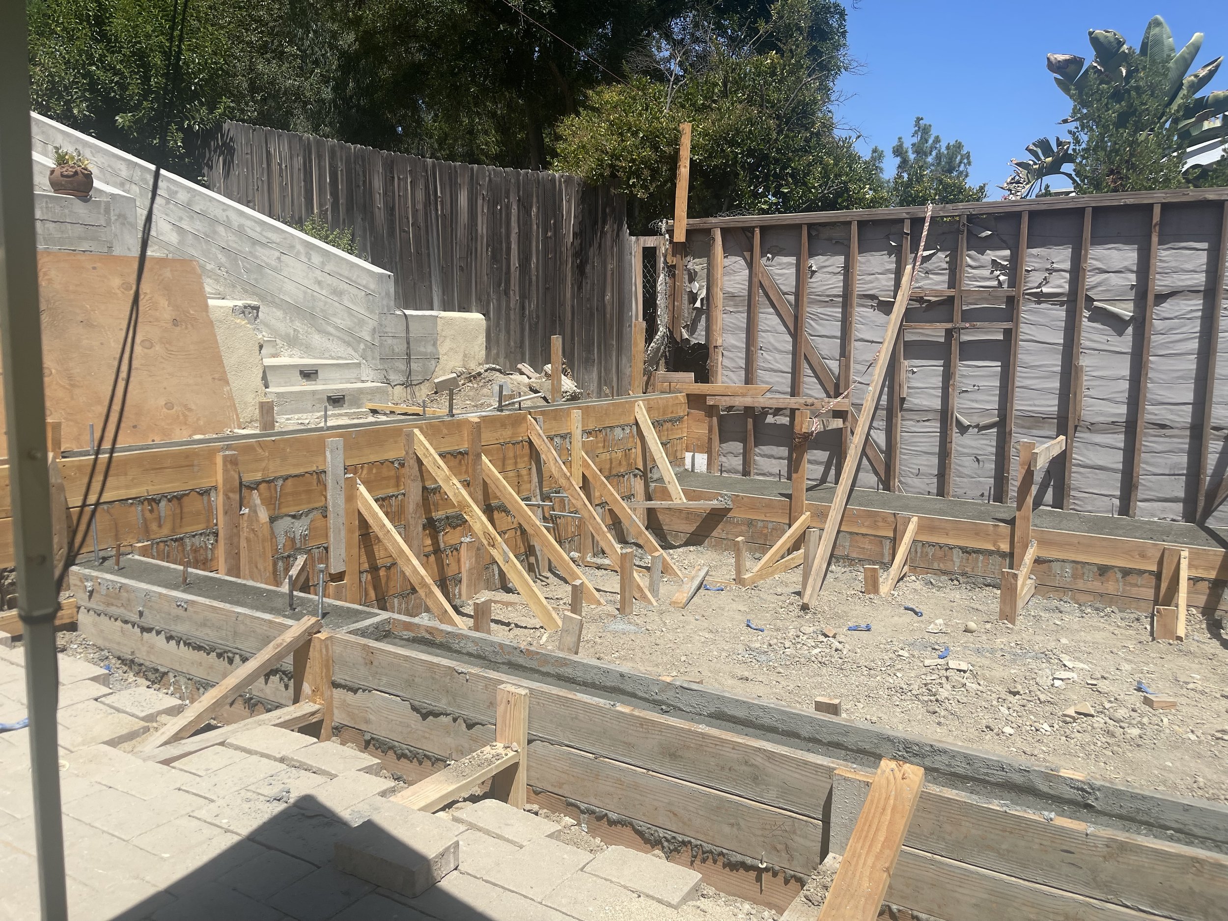 Construction site with wooden framework, partially built concrete steps, and a wooden fence in the background, under a clear blue sky.
