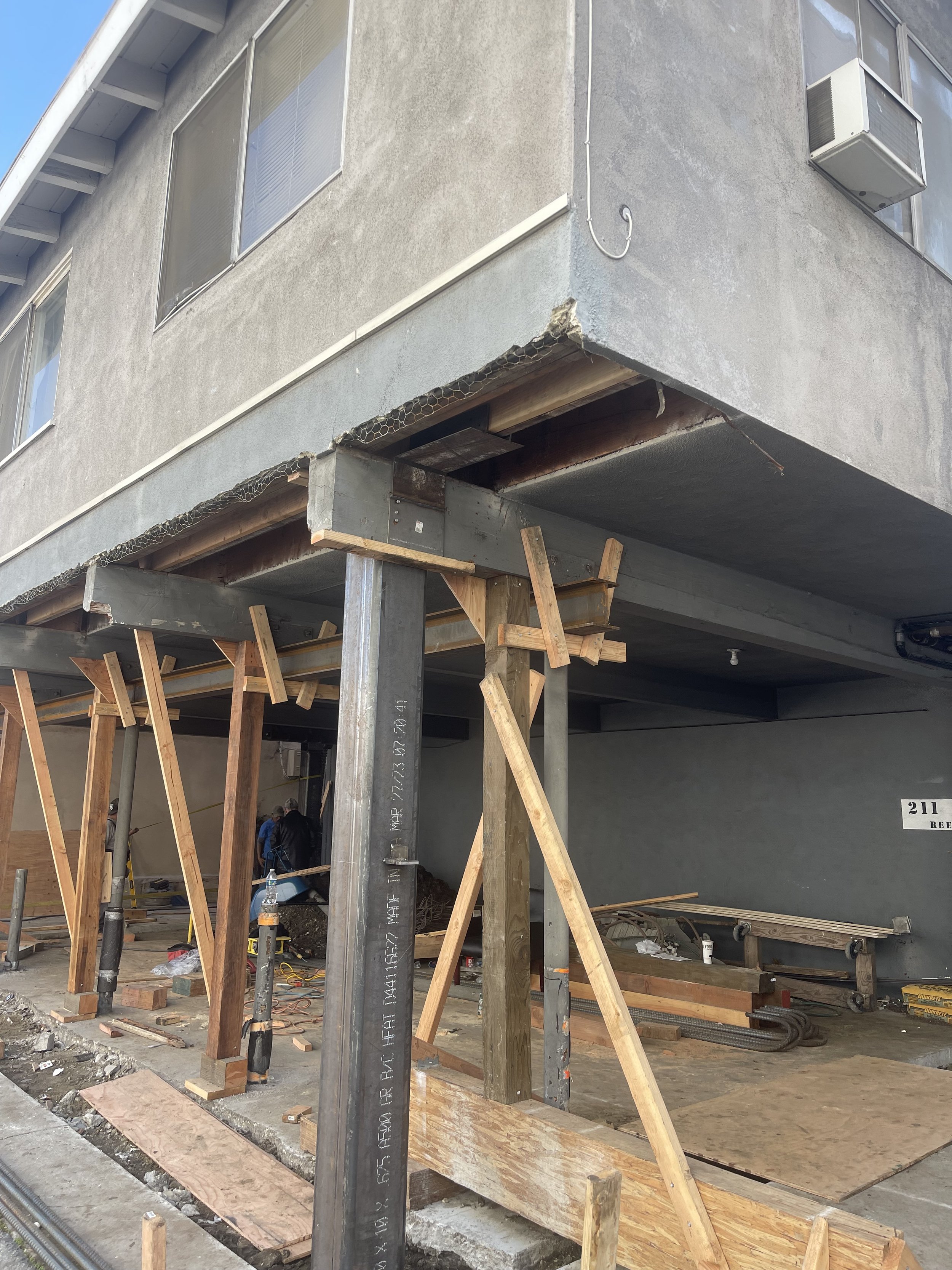 Construction site showing the support structure for a building's extension, with wooden beams and metal posts under a concrete balcony.