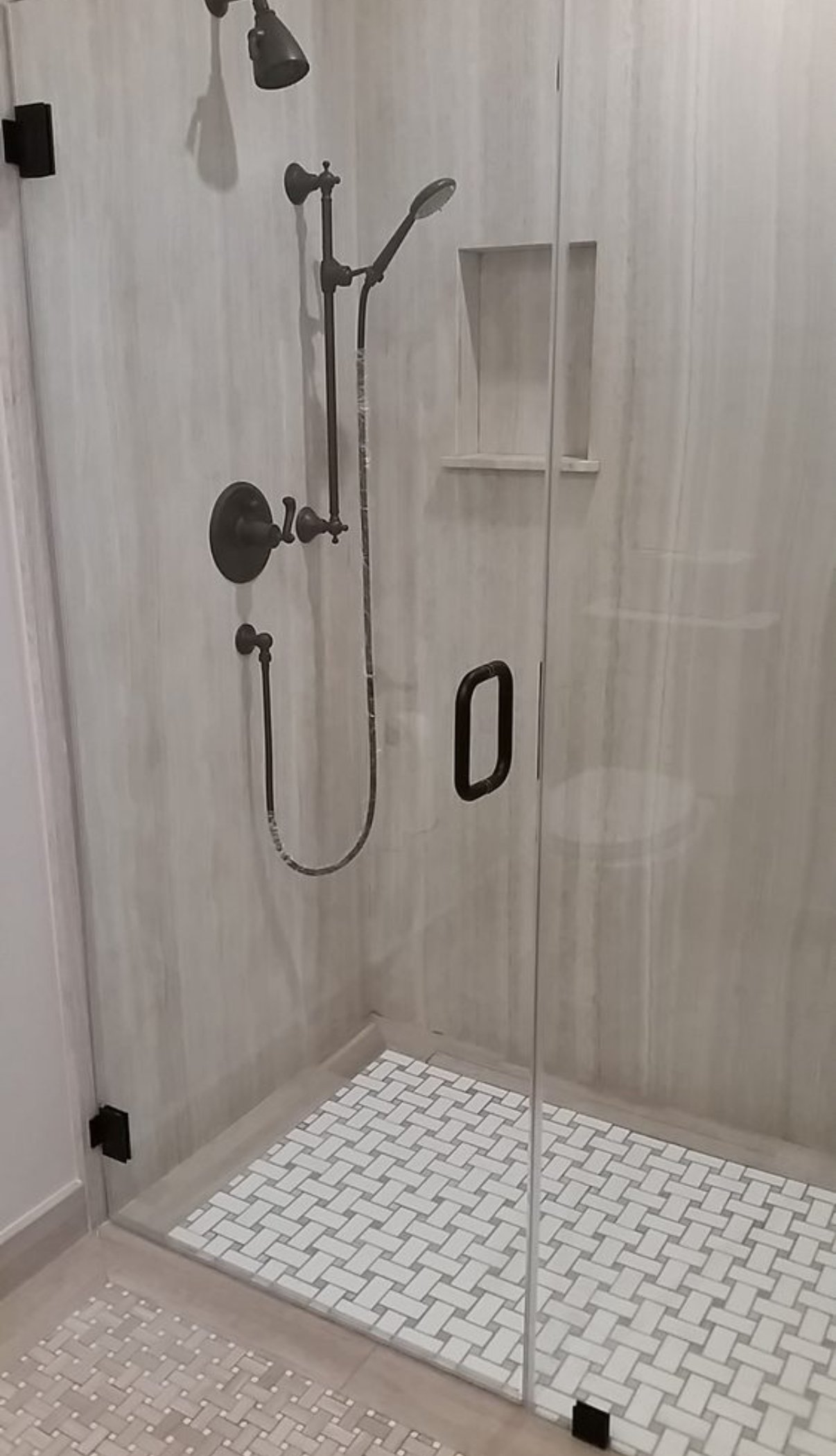 A walk-in shower with a glass door, black fixtures, and a built-in wall shelf, with a tile floor and light-colored walls.