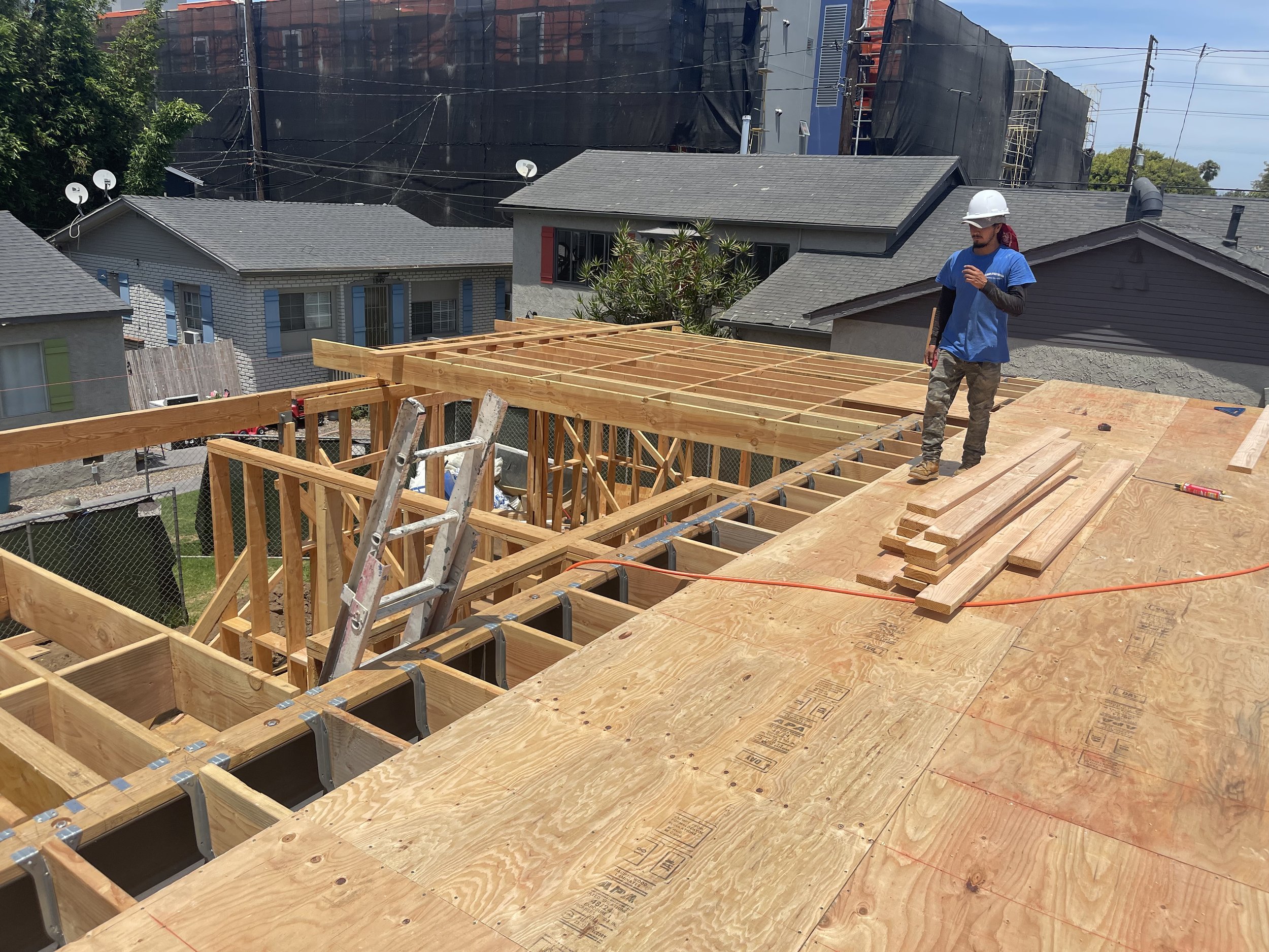 Construction worker wearing a white safety helmet and blue shirt standing on a building under construction, with wooden beams and plywood sheets. There are houses and a building with black netting in the background.
