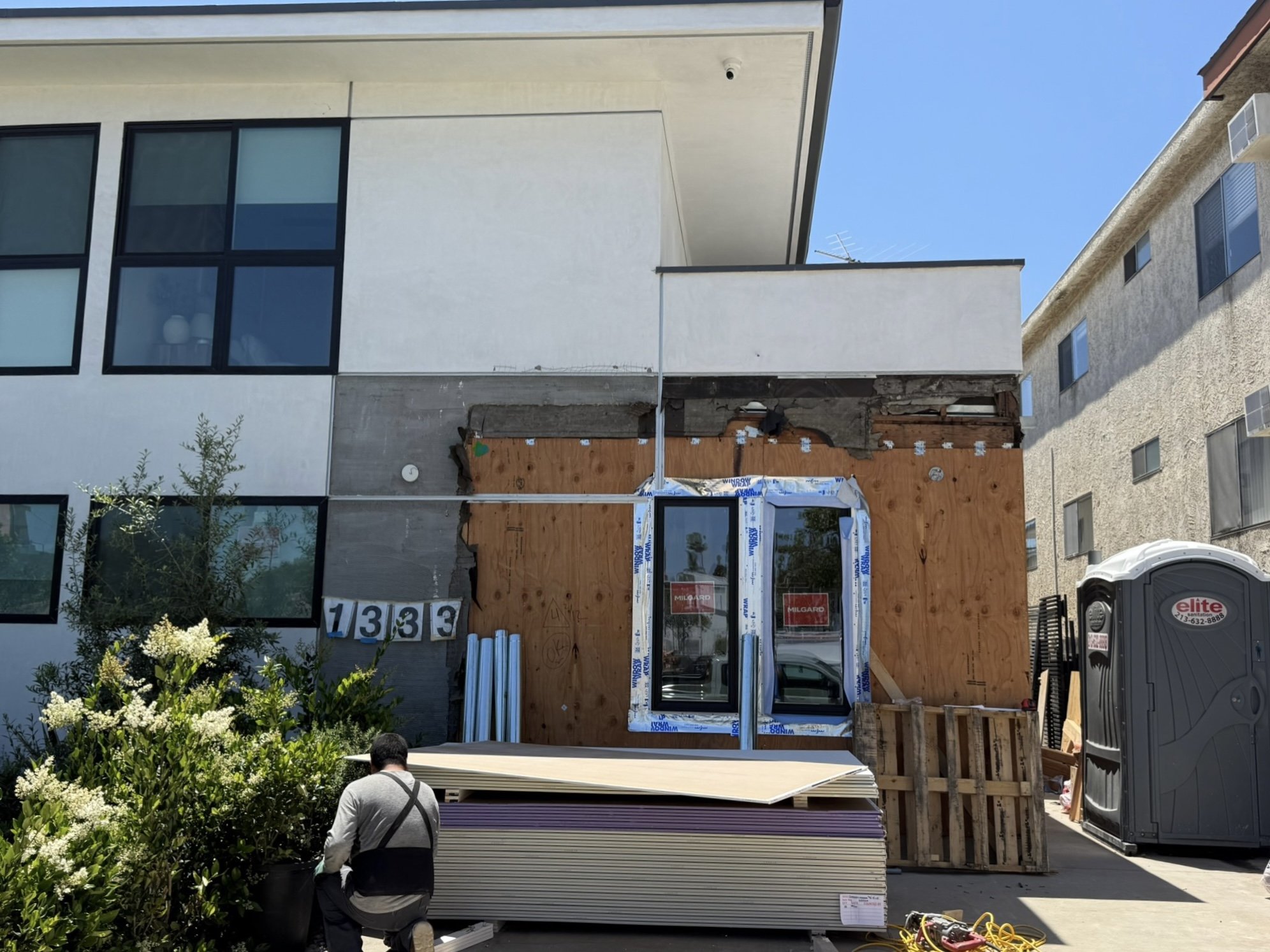 Construction site with a man kneeling near plants, plywood, and building materials in front of a house with an attached porch, during daytime with clear blue sky.