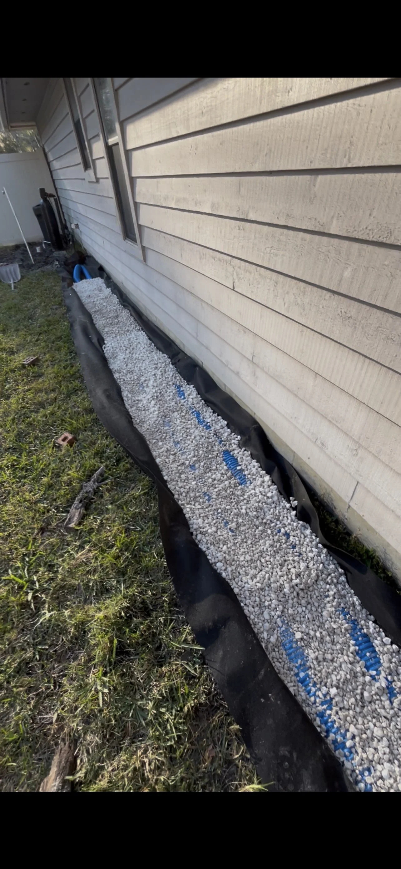 Heart-shaped garden bed with white gravel, black landscaping fabric, and blue landscape edging along a beige house exterior.