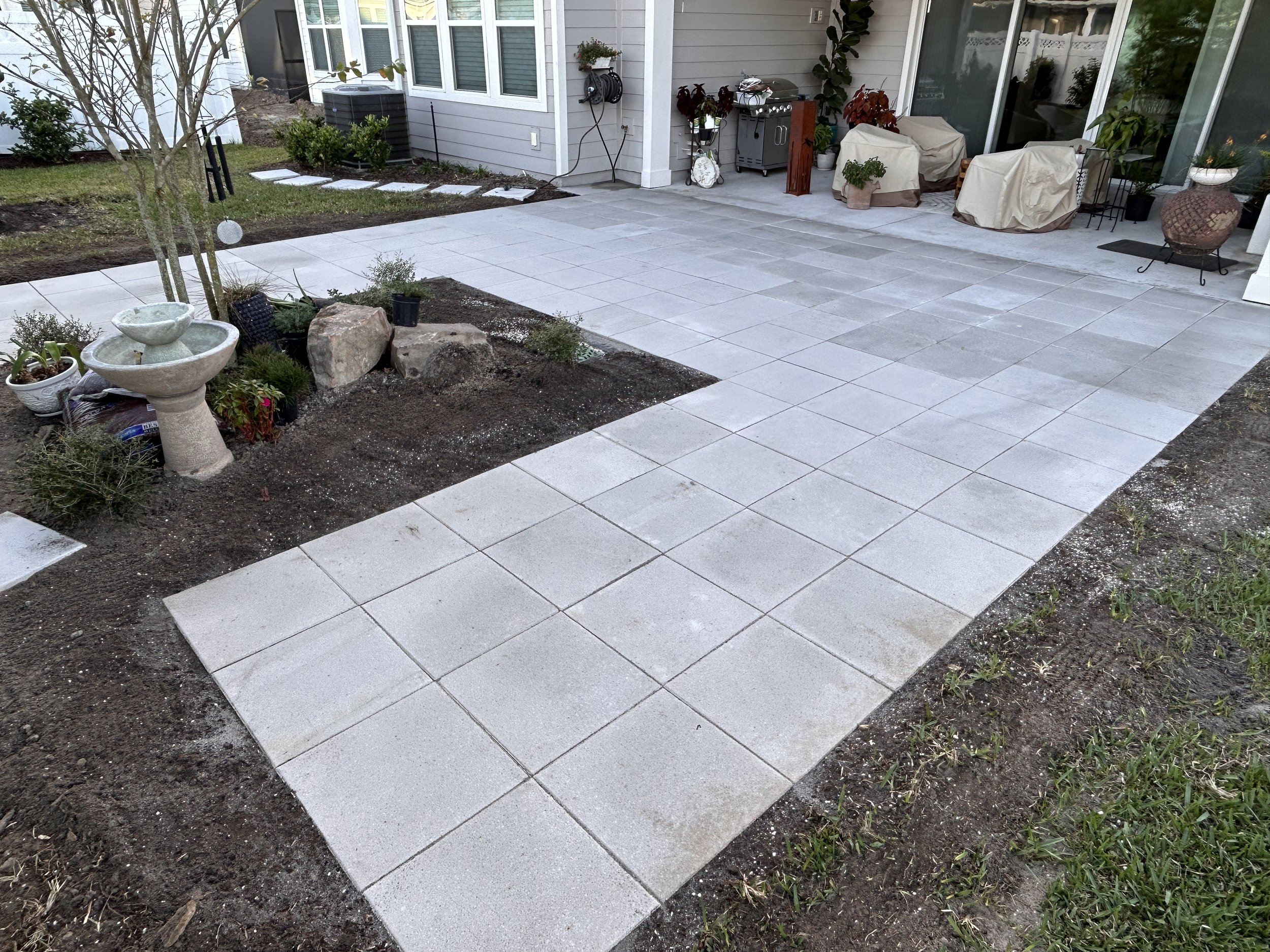 Backyard patio with newly laid concrete pavers, garden area with rocks, plants, and a birdbath, and patio furniture covered with beige covers near the house.