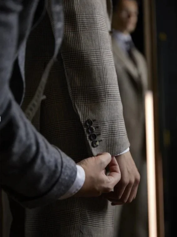 A person adjusting the sleeve of a brown checkered blazer while standing indoors, with another person blurred in the background.