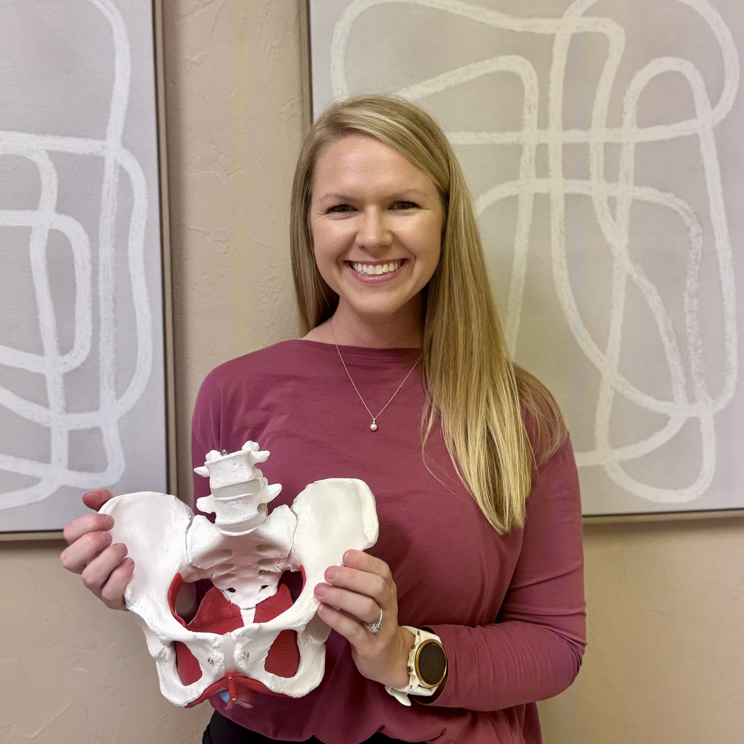 A pelvic floor therapist with long blonde hair, wearing a pink long sleeve shirt, holding a white pelvic skeleton model, smiling, standing in front of abstract line art on the wall.