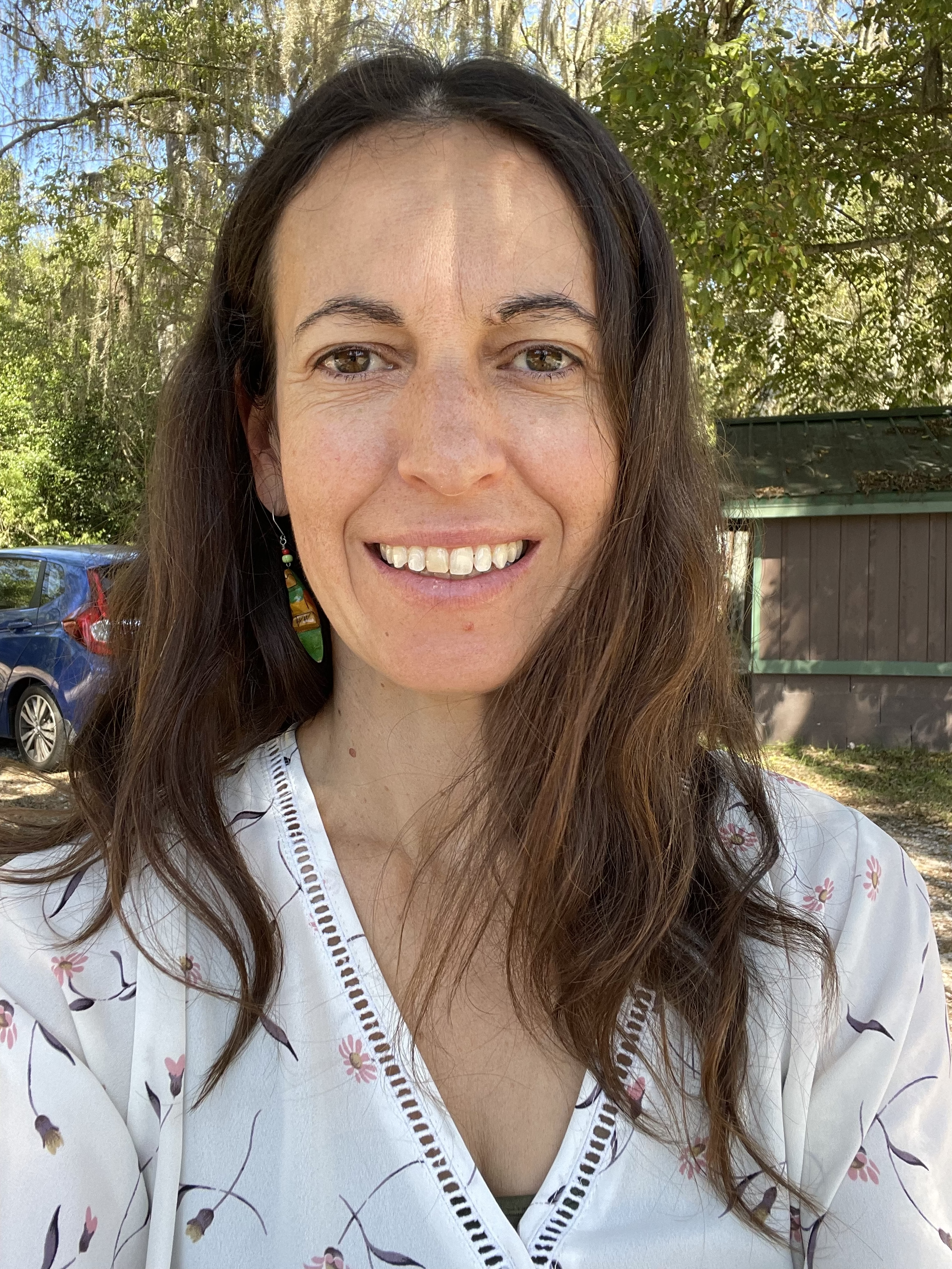 A woman with long brown hair smiling outdoors, wearing a white top with floral pattern and colorful earrings, with trees and a car in the background.