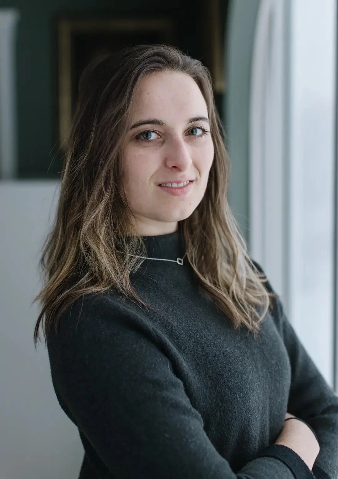 A young woman with long, wavy brown hair and blue eyes standing near a window, wearing a black top and a silver necklace, smiling slightly at the camera.