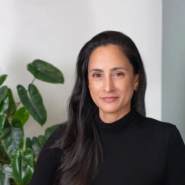 headshot of a woman with long dark hair smiling at the camera with green plants in the background