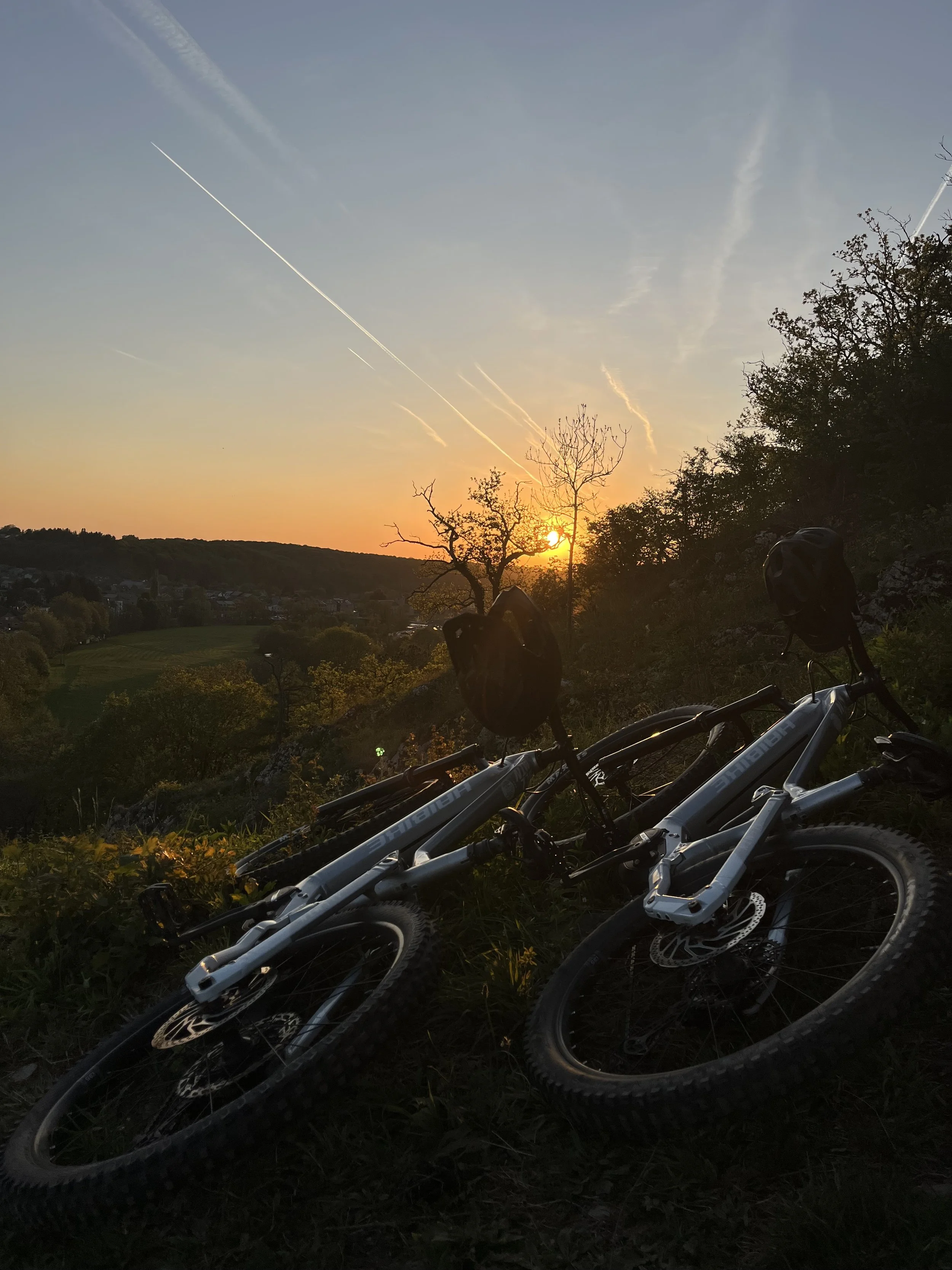 Cinq vélos électriques reposent sur le sol en contrebas d'une colline lors d'un coucher de soleil avec un ciel teinté d'orange et de bleu, entouré de végétation et de quelques arbres nus.