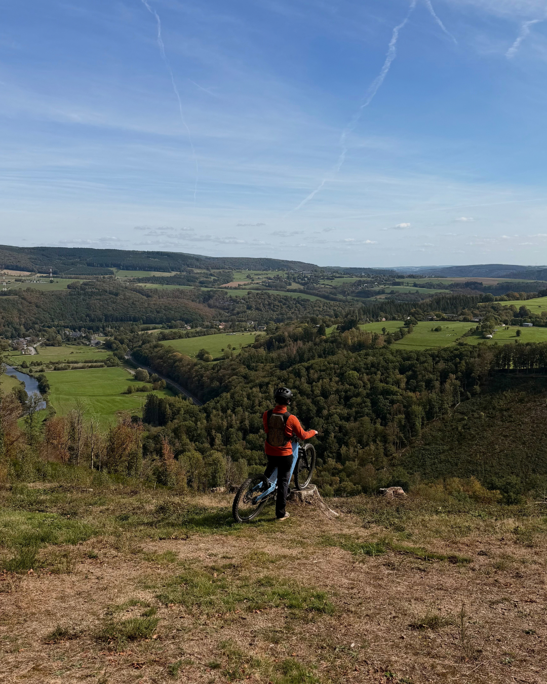 Personne en VTT regardant un paysage vallonné avec une rivière, champs et arbres sous un ciel bleu avec des cirrus.